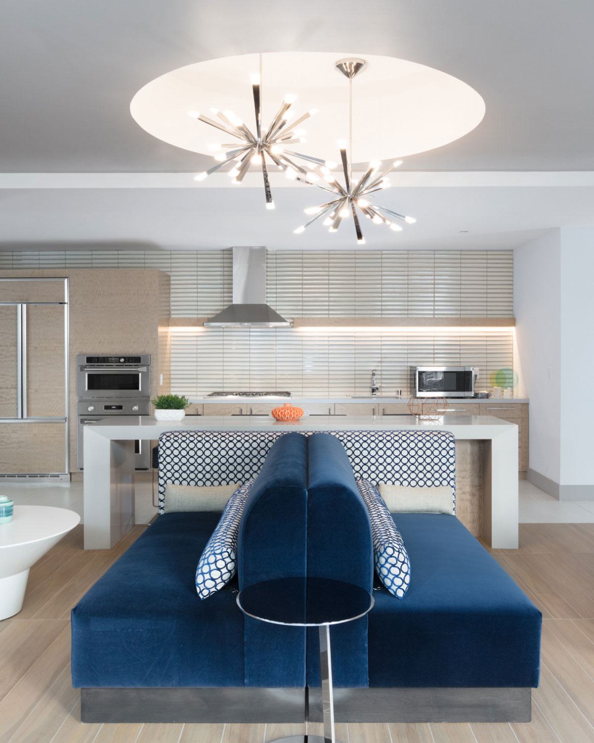 Modern apartment interior at The Triton featuring a sleek open kitchen with stainless steel appliances, light wood cabinetry, and a stylish navy blue sectional sofa beneath contemporary starburst pendant lighting.