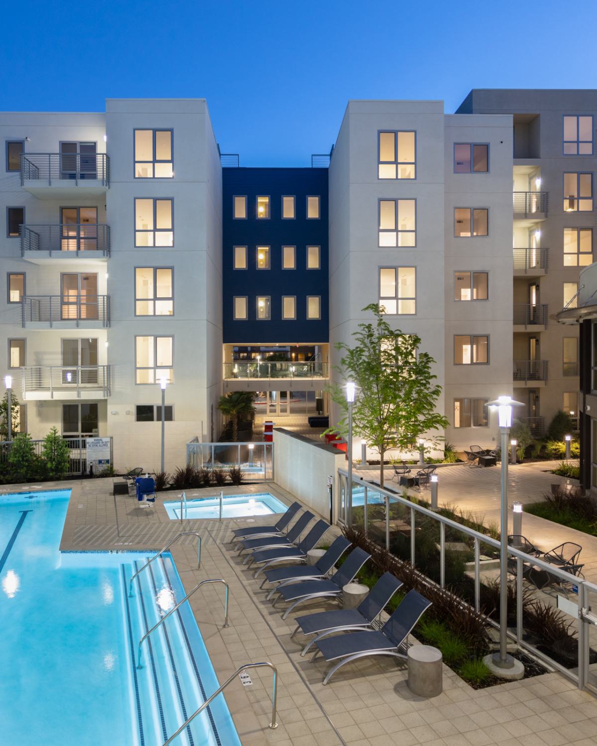 Evening view of The Triton Apartments courtyard with a lit swimming pool, lounge chairs, hot tubs, and modern apartment buildings surrounding the space.