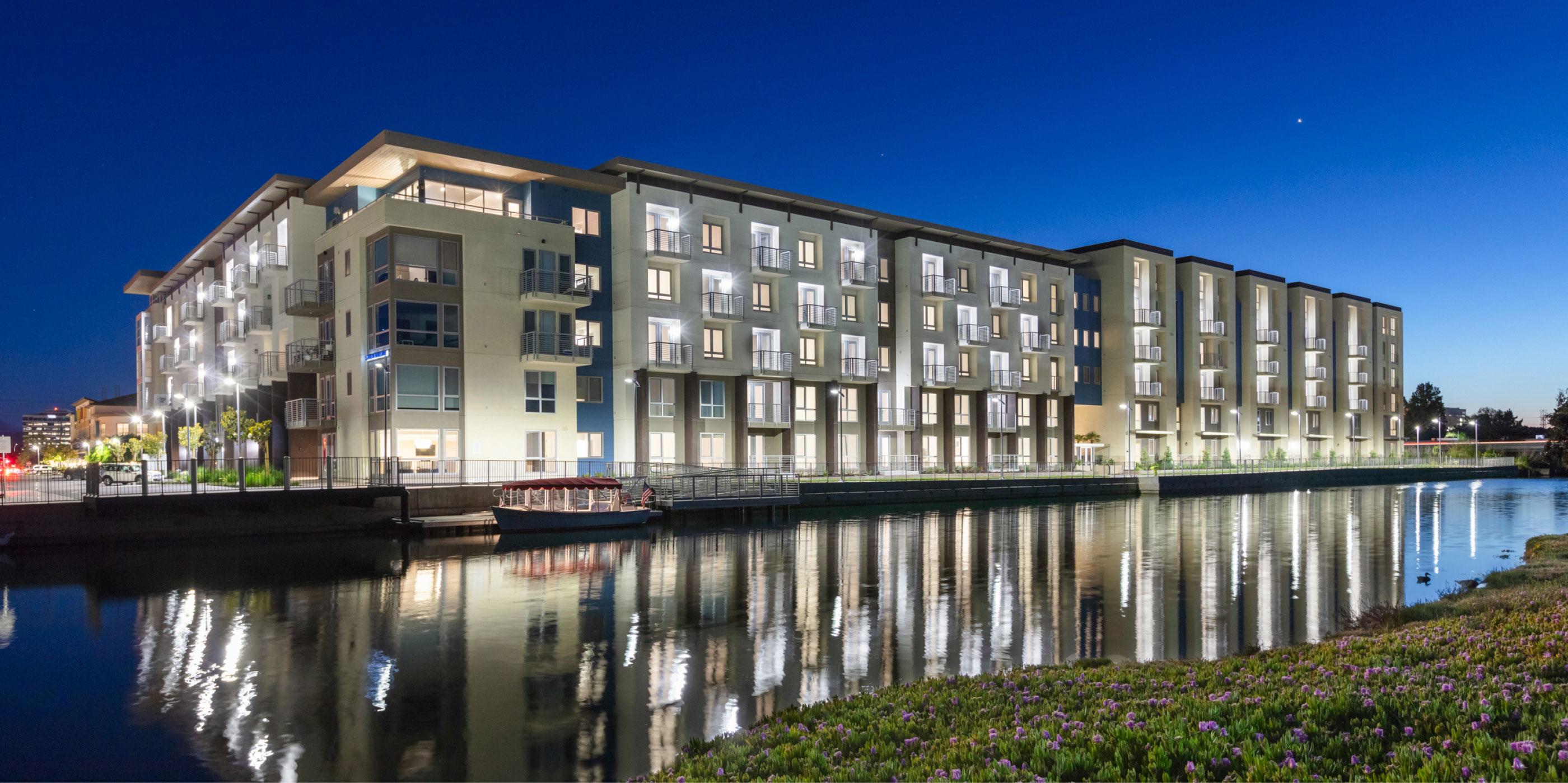 Nighttime exterior of The Triton Apartments in Foster City with modern waterfront buildings illuminated and reflecting across the calm lagoon, with landscaped greenery in the foreground.