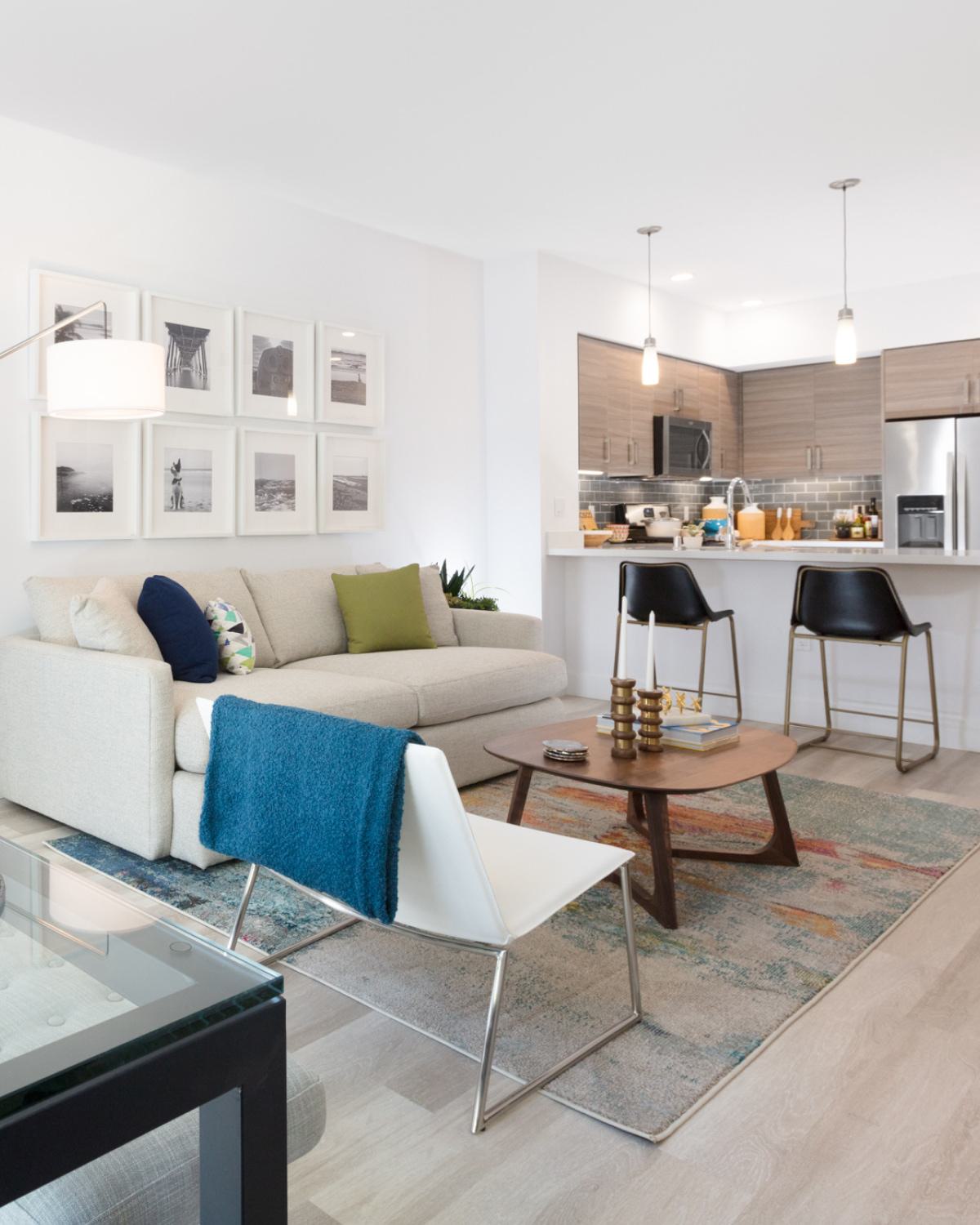 Bright modern apartment living room at The Triton featuring a neutral sofa with colorful pillows, open kitchen with stainless steel appliances, dining area with glass table, and contemporary decor accents.