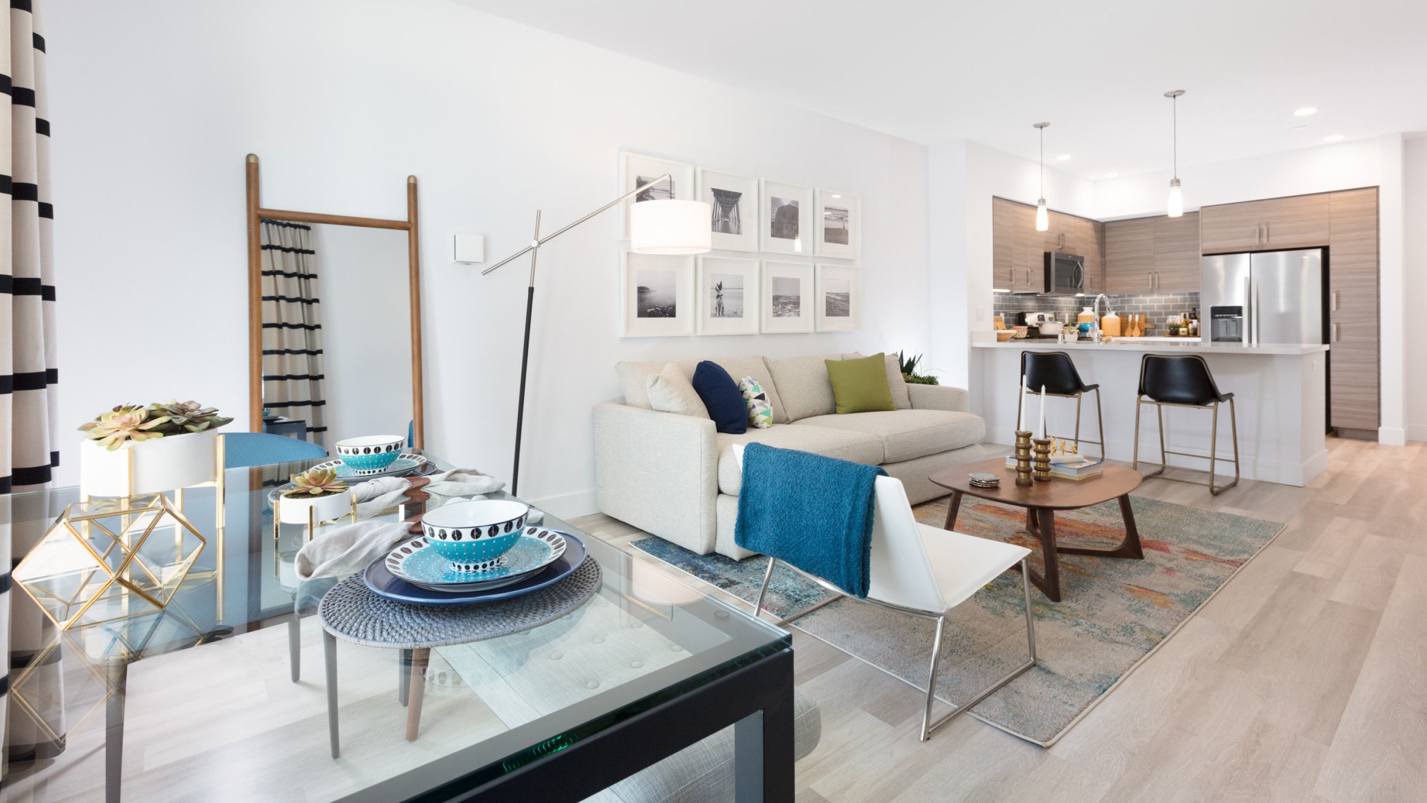 Bright modern apartment living room at The Triton featuring a neutral sofa with colorful pillows, open kitchen with stainless steel appliances, dining area with glass table, and contemporary decor accents.