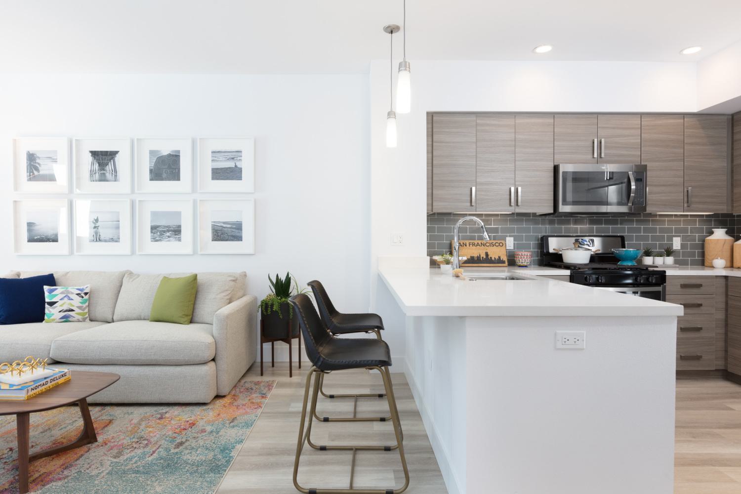 Bright modern apartment living room at The Triton featuring a neutral sofa with colorful pillows, open kitchen with stainless steel appliances, dining area with glass table, and contemporary decor accents.