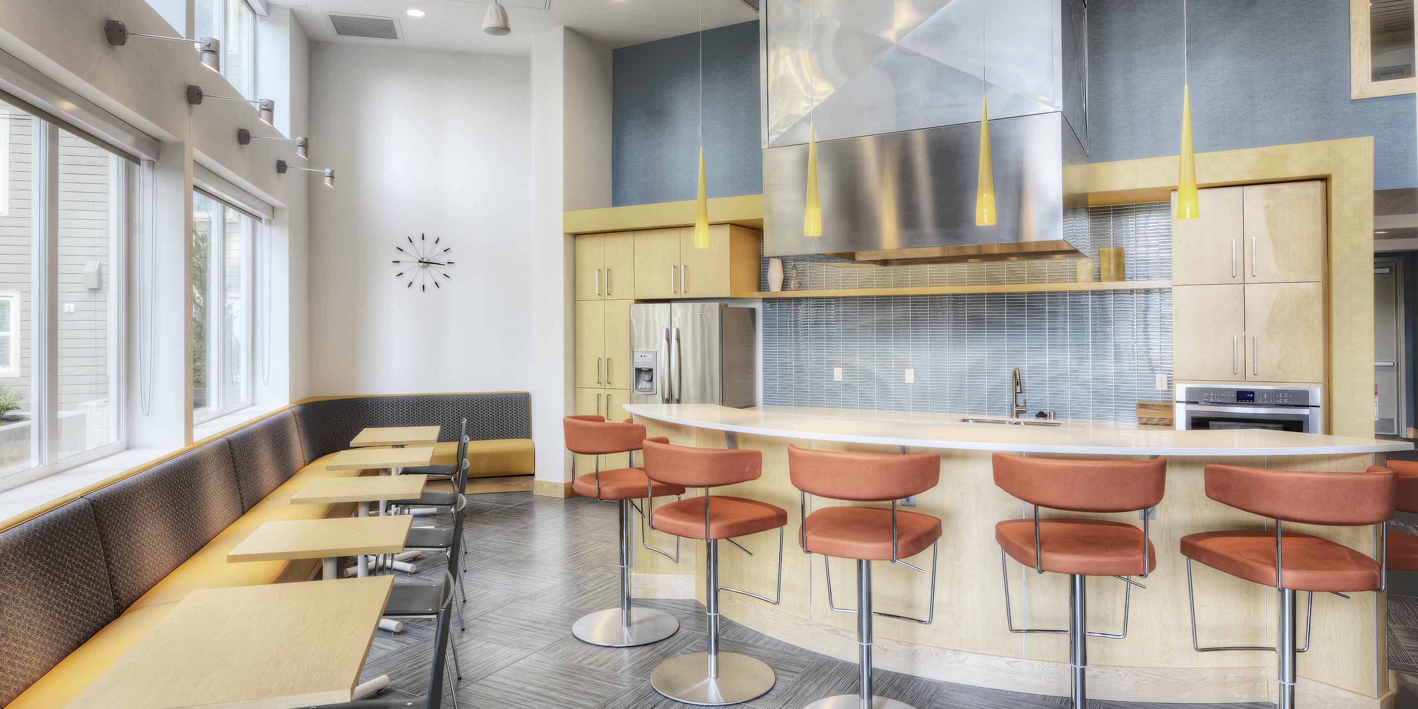 Bright community kitchen and dining area featuring a modern island with barstools, stainless steel appliances, and ample seating along the windows.