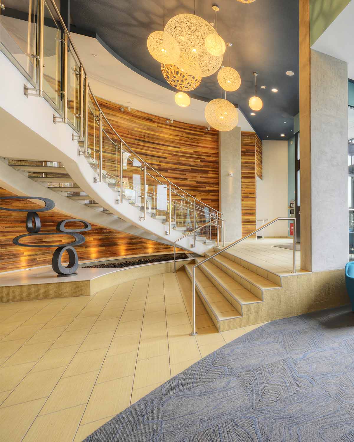 A modern apartment lobby in Ballard, WA, featuring a curved staircase, wood accent wall, pendant lights, and cozy seating by large street-facing windows