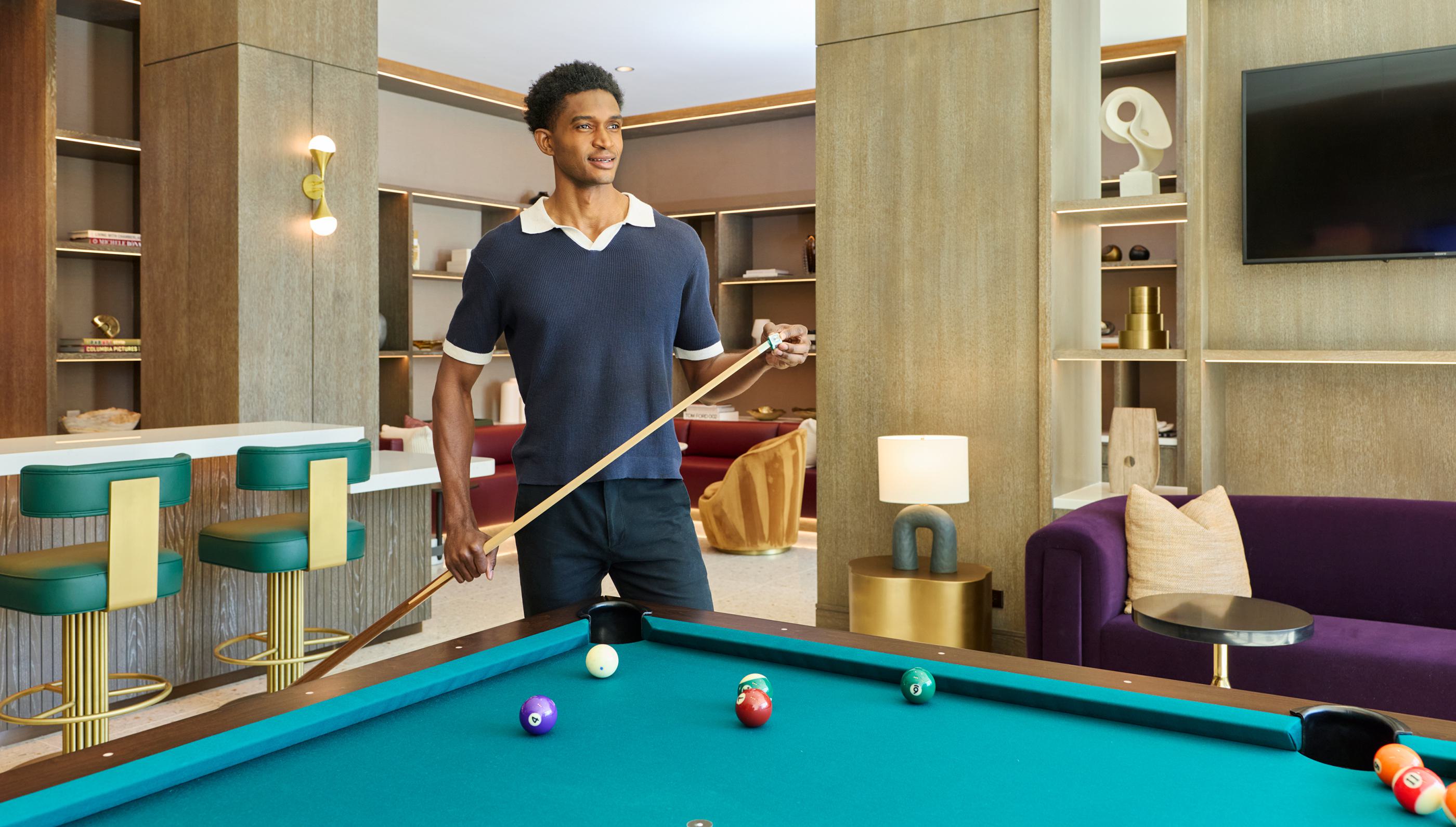 stylish game and media lounge featuring a central pool table, wall-mounted TVs displaying a baseball game, plush seating, and built-in shelving.