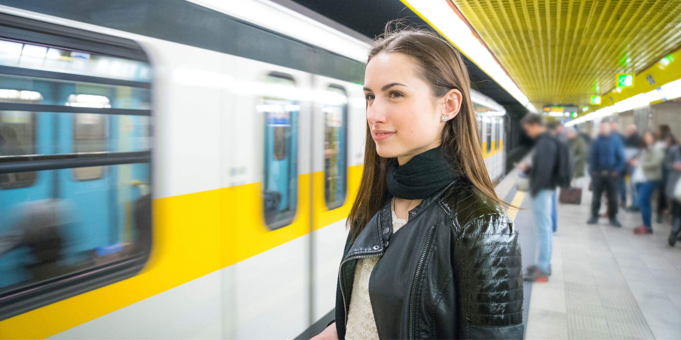 Young woman waiting at an underground metro station with a yellow-striped train arriving, wearing a black leather jacket and scarf, with commuters in the background.