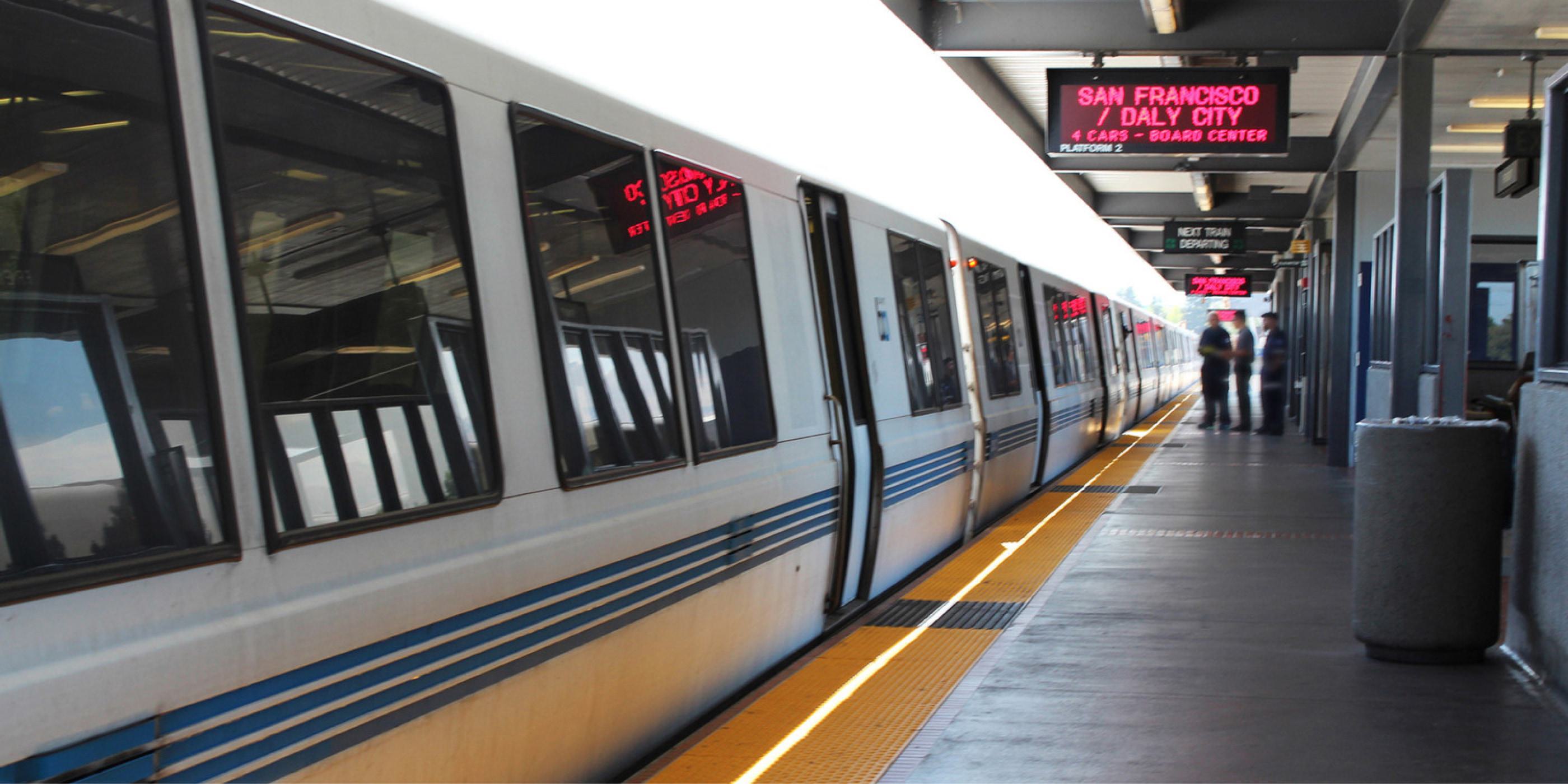 Young woman waiting at an underground metro station with a yellow-striped train arriving, wearing a black leather jacket and scarf, with commuters in the background.