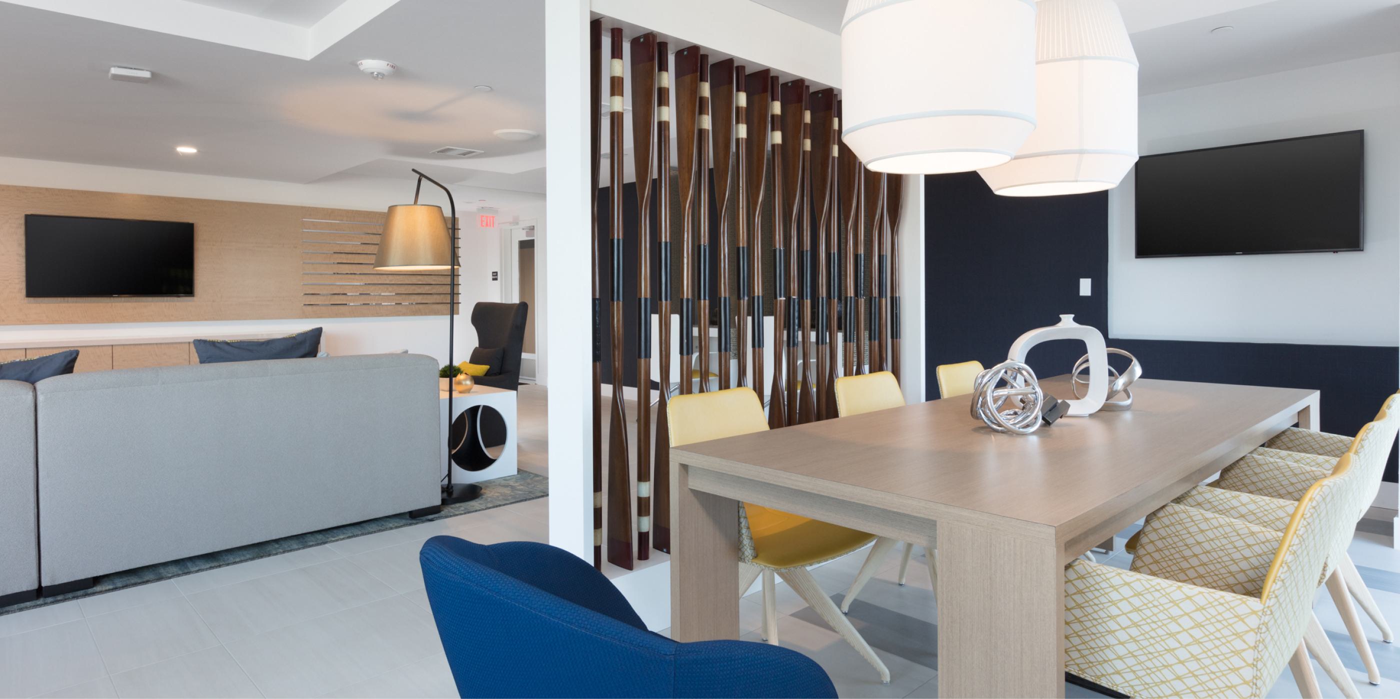 Contemporary lounge and dining area at The Triton Apartments featuring a light wood table with colorful chairs, sculptural decor, modern pendant lighting, and a partial view of a cozy seating area divided by vertical wood accents.