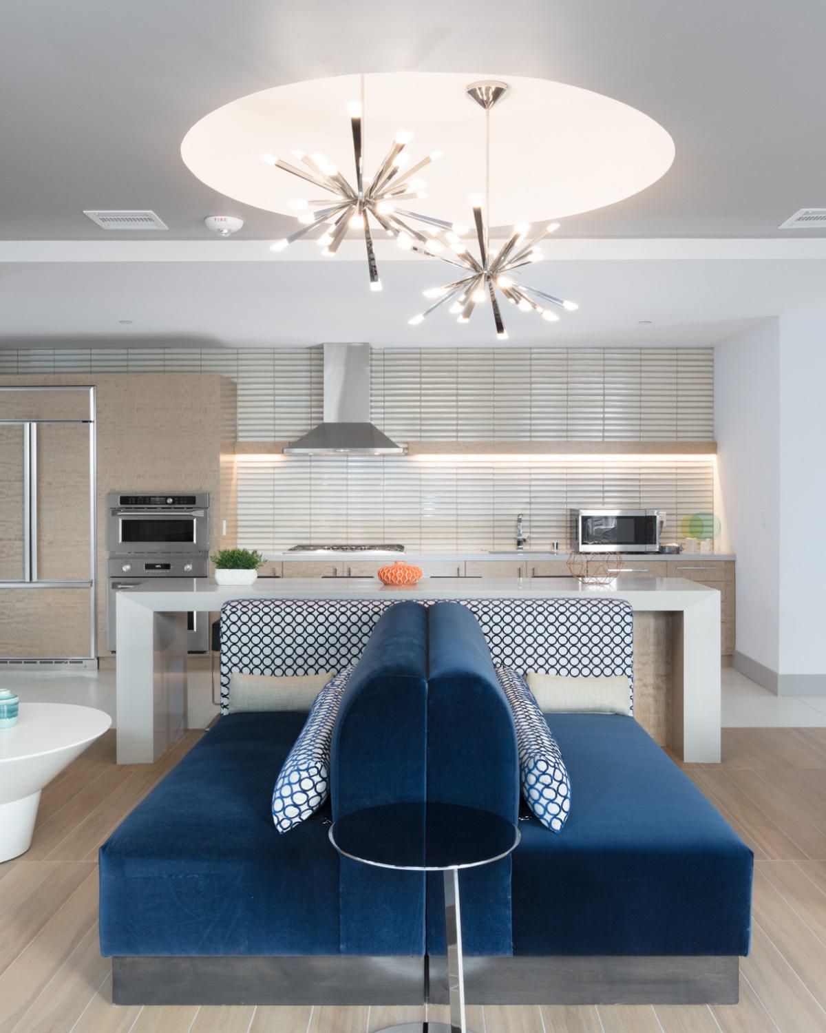 Sleek business lounge at The Triton Apartments featuring a modern kitchen with stainless steel appliances, a cozy fireplace, stylish blue seating, and contemporary lighting.