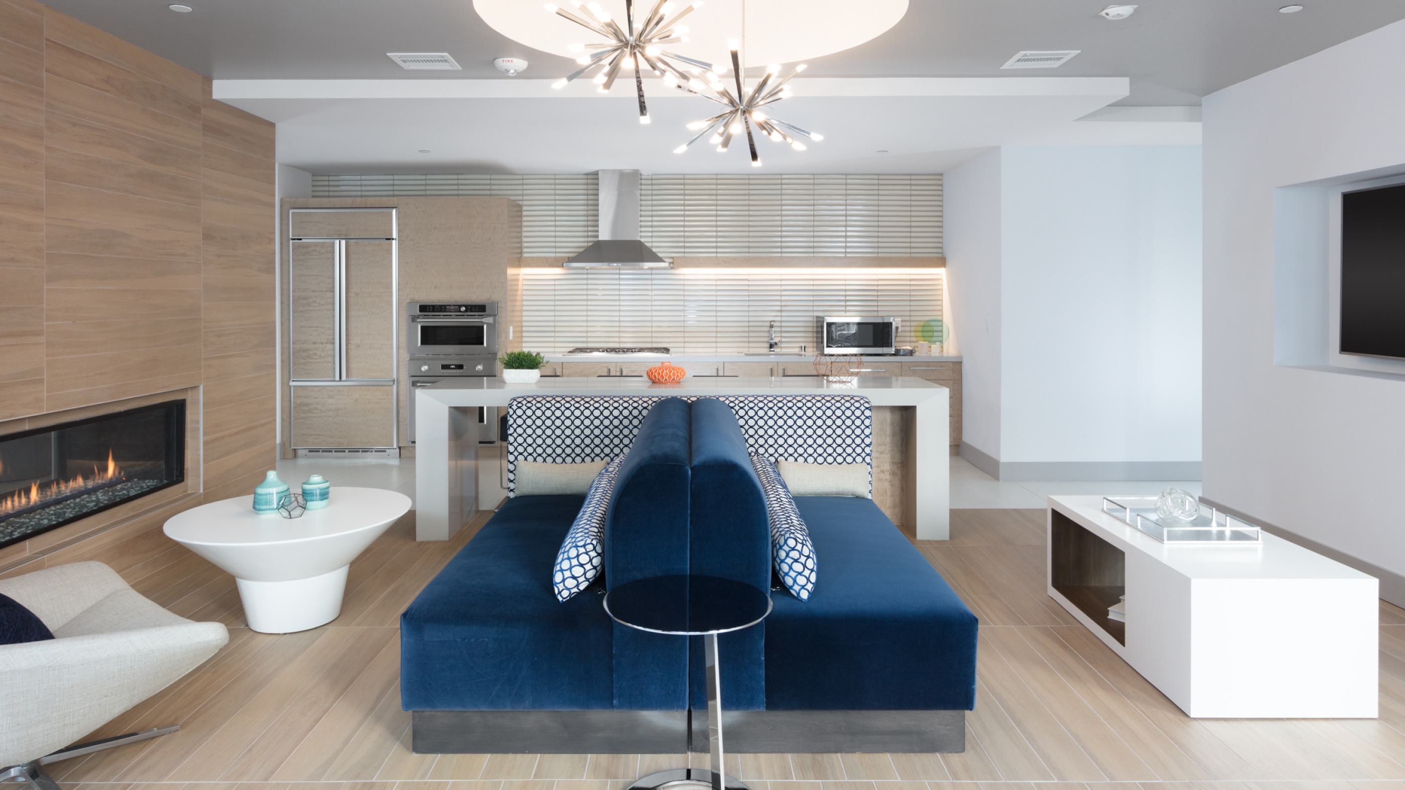 Sleek business lounge at The Triton Apartments featuring a modern kitchen with stainless steel appliances, a cozy fireplace, stylish blue seating, and contemporary lighting.