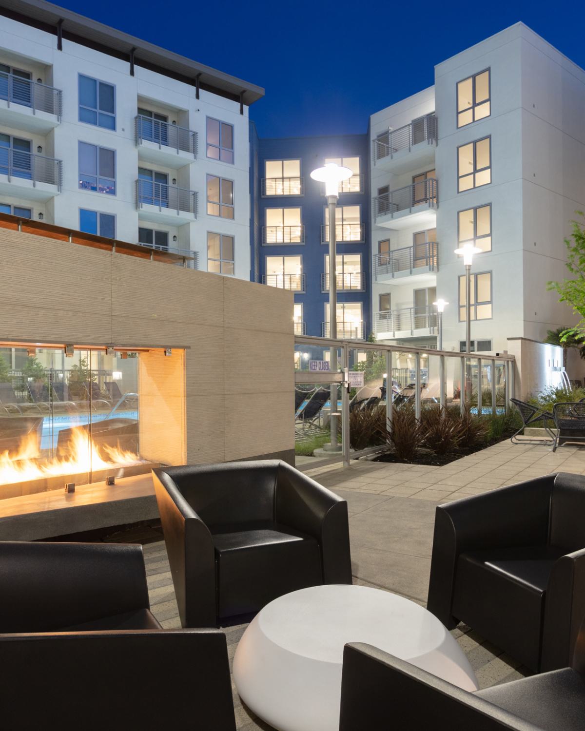 Evening courtyard at The Triton Apartments featuring a modern outdoor fireplace with glass enclosure, black lounge chairs, and surrounding apartment buildings with illuminated windows.
