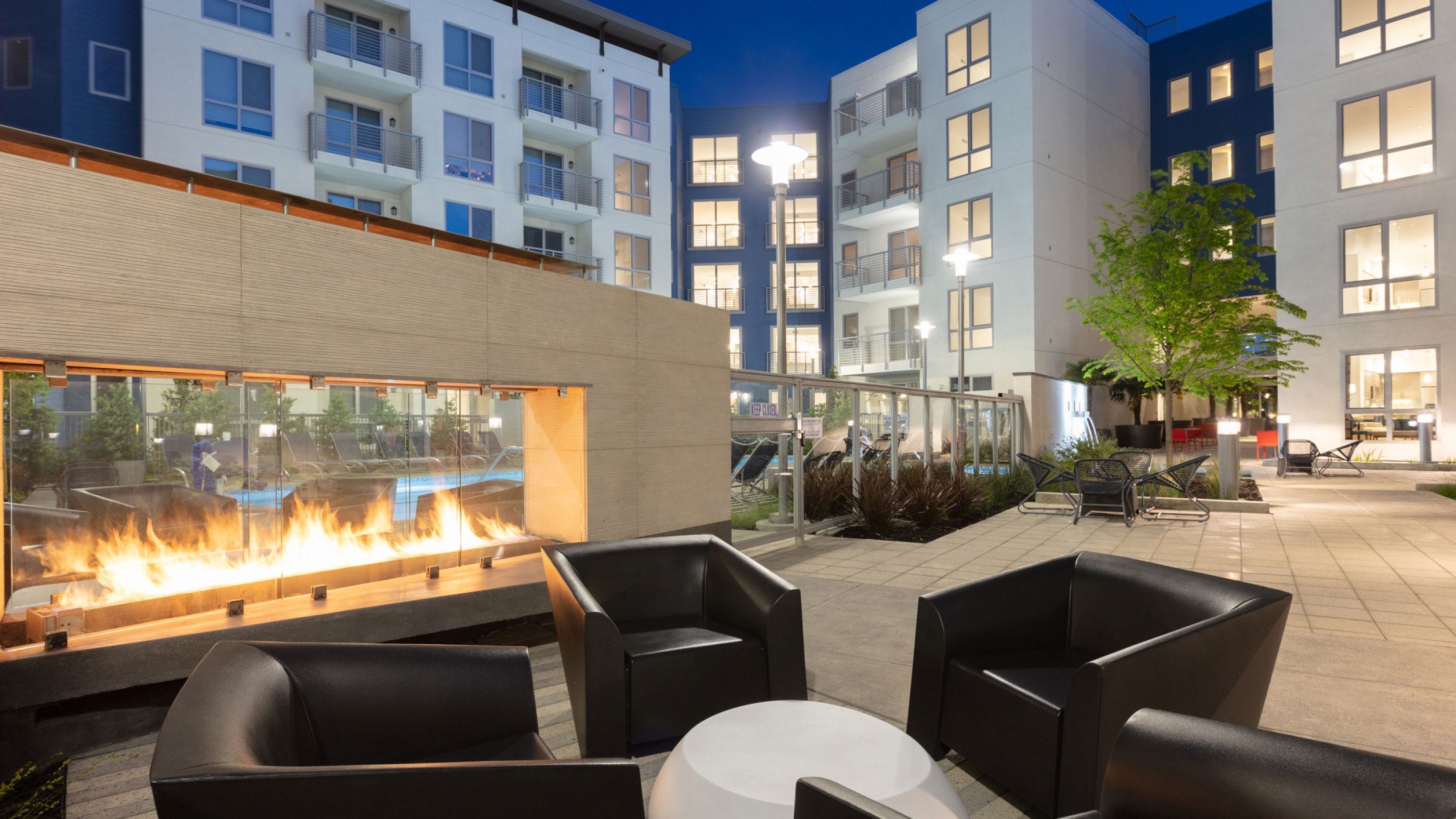 Evening courtyard at The Triton Apartments featuring a modern outdoor fireplace with glass enclosure, black lounge chairs, and surrounding apartment buildings with illuminated windows.