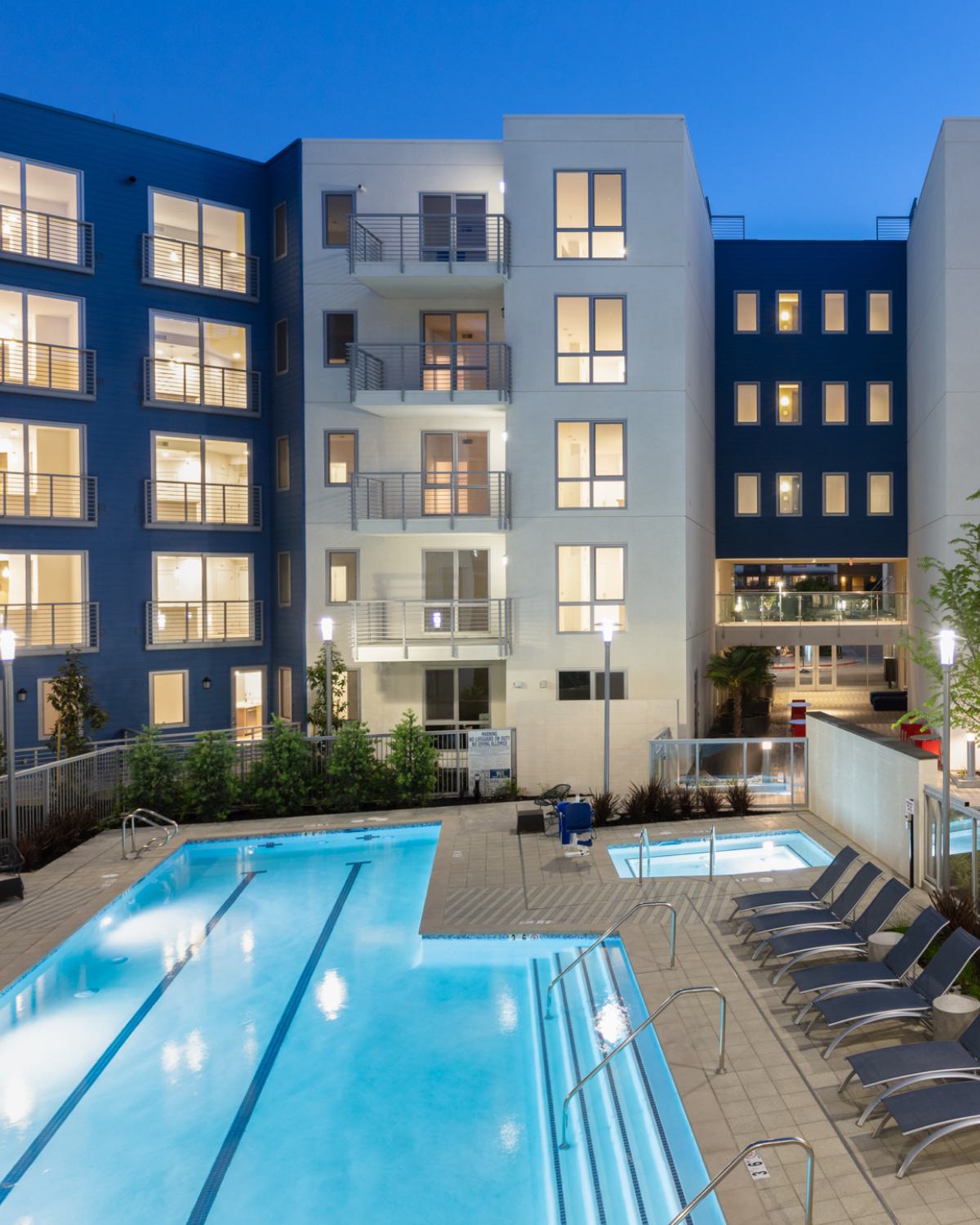 Evening view of The Triton Apartments courtyard with a lit swimming pool, hot tubs, lounge chairs, and modern apartment buildings surrounding the space.