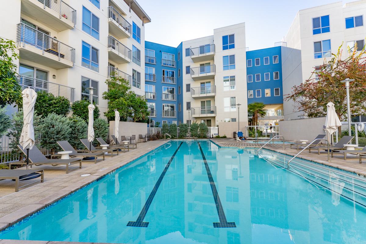 View of The Triton Apartments courtyard with a lit swimming pool, hot tubs, lounge chairs, and modern apartment buildings surrounding the space.