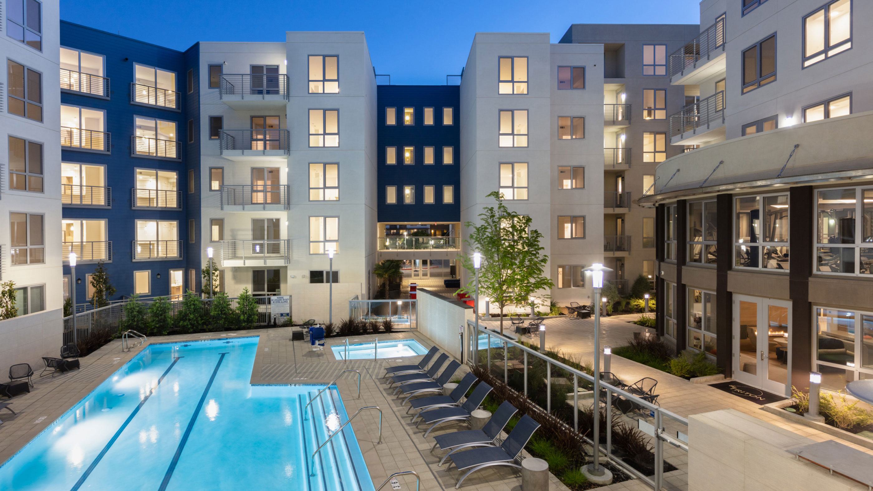 Evening view of The Triton Apartments courtyard with a lit swimming pool, hot tubs, lounge chairs, and modern apartment buildings surrounding the space.