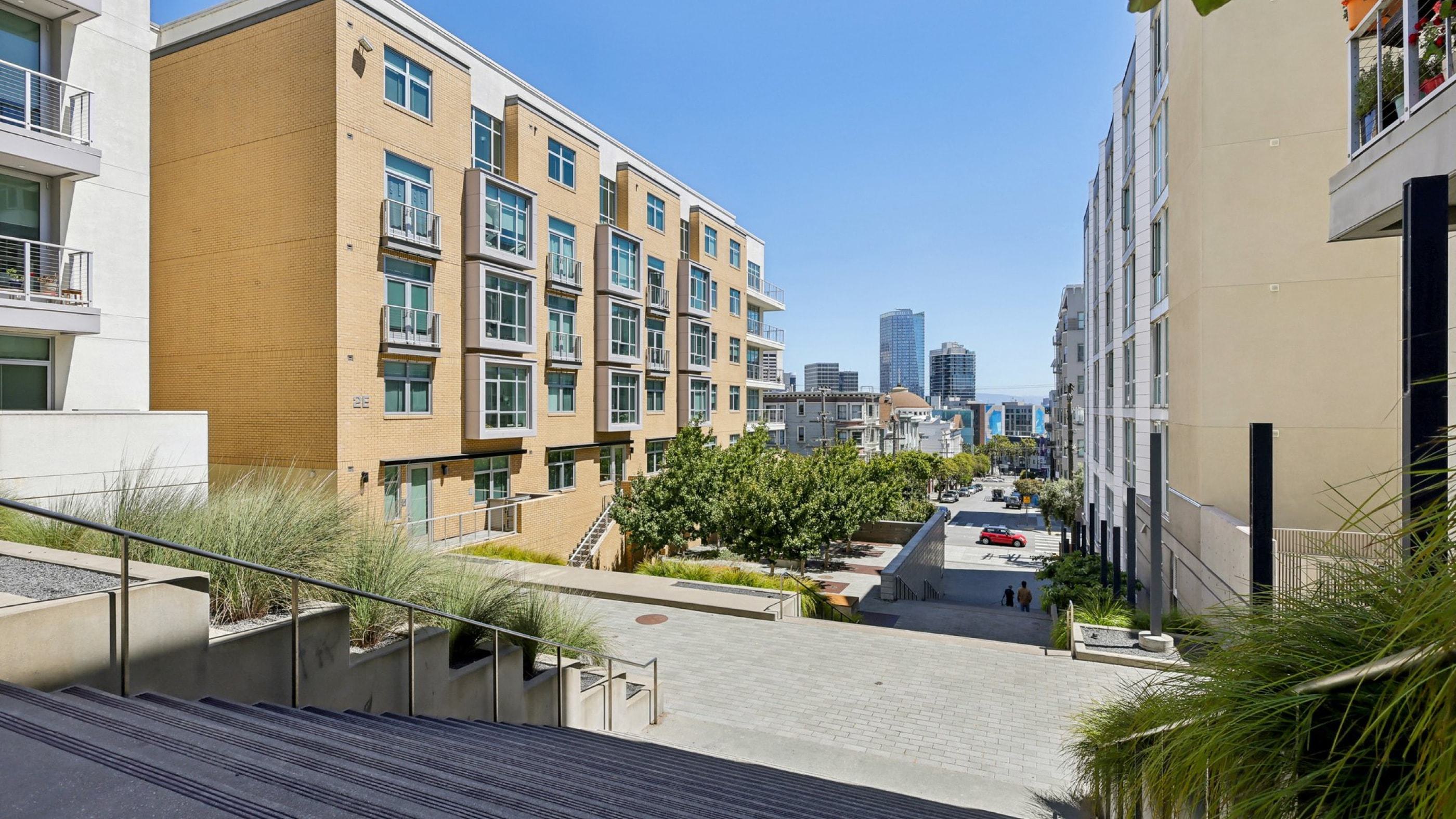 Alchemy modern mid-rise apartment buildings with a terraced walkway framed by greenery that leads down toward the city, offering peekaboo views of downtown high-rises and a glimpse of the San Francisco bay beyond.