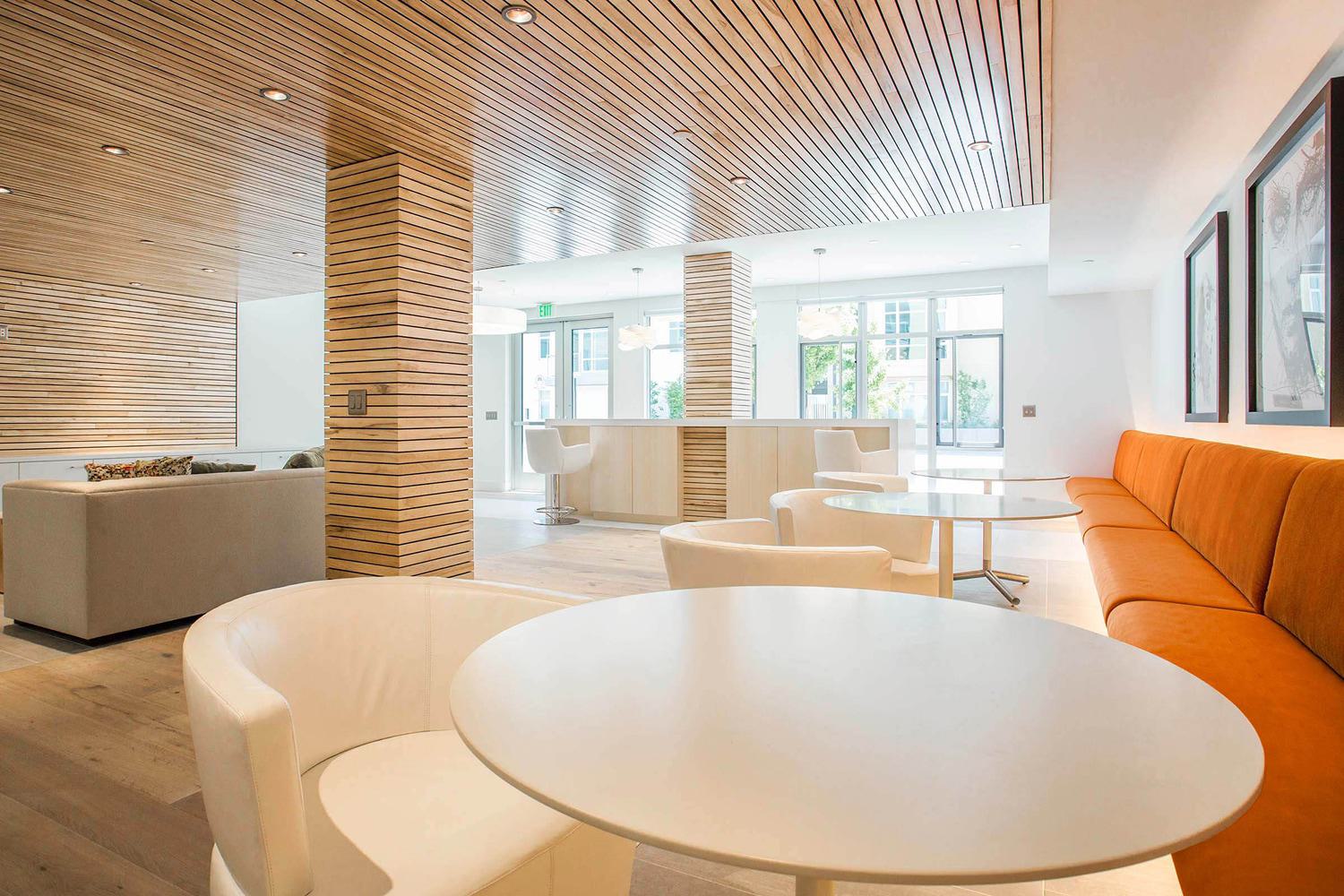 This bright, inviting lounge features clean white tables and chairs, a long orange banquette, and warm wood slat ceilings and columns. Natural light floods the space through large windows, creating a modern yet welcoming atmosphere ideal for casual gatherings or work sessions.