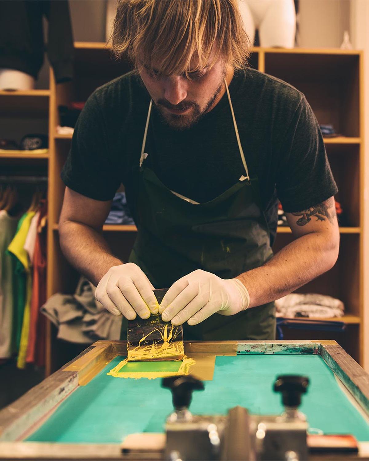 A focused artist in a green apron and gloves carefully screen prints a design with vibrant yellow ink. The scene captures a hands-on creative moment, surrounded by colorful garments and studio shelving in the background.