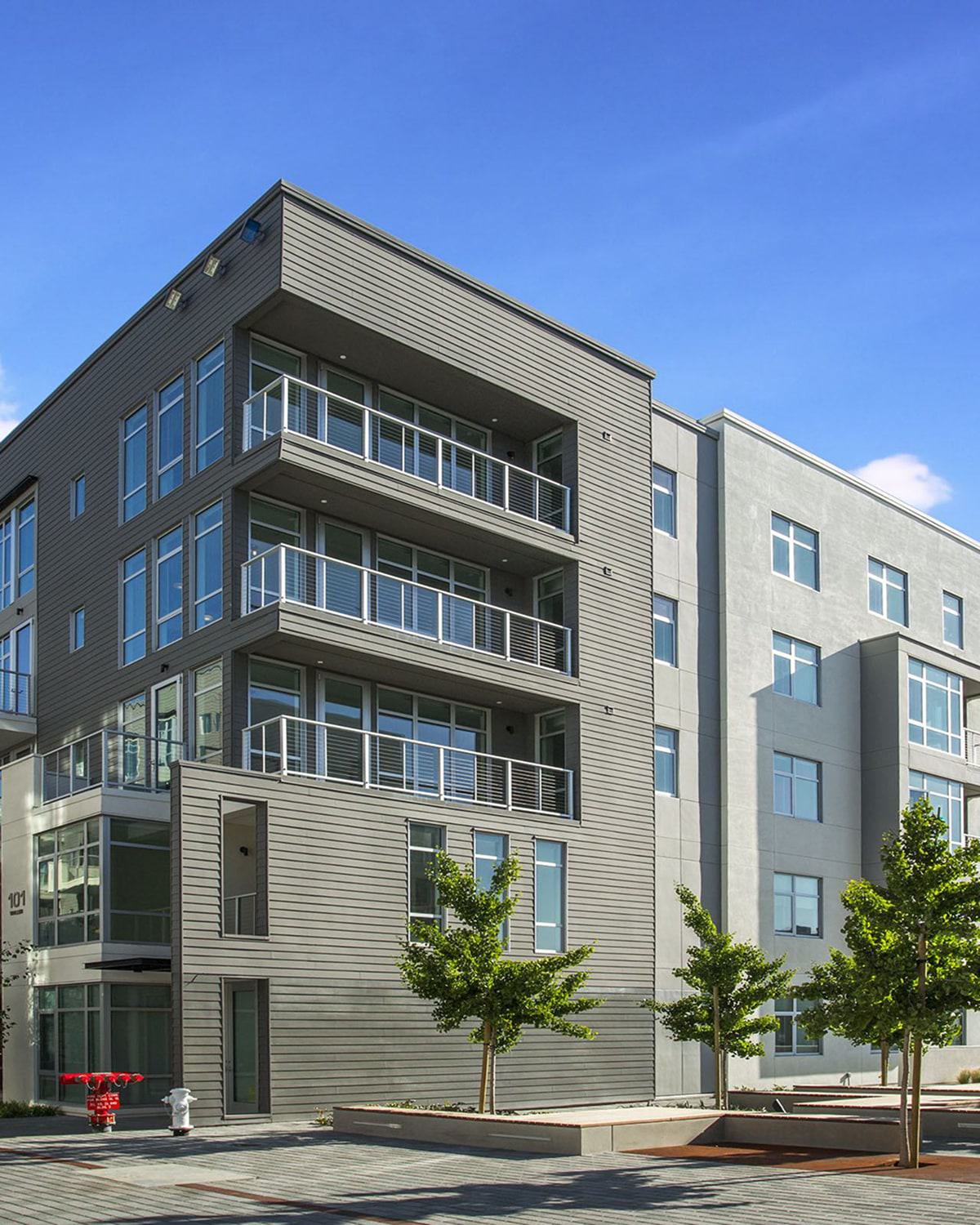 Alchemy contemporary apartment building in Hayes Valley with clean lines, large windows, and private balconies, framed by sleek siding and light neutral tones. Young trees line the paved courtyard below, giving the space a modern, walkable neighborhood feel.