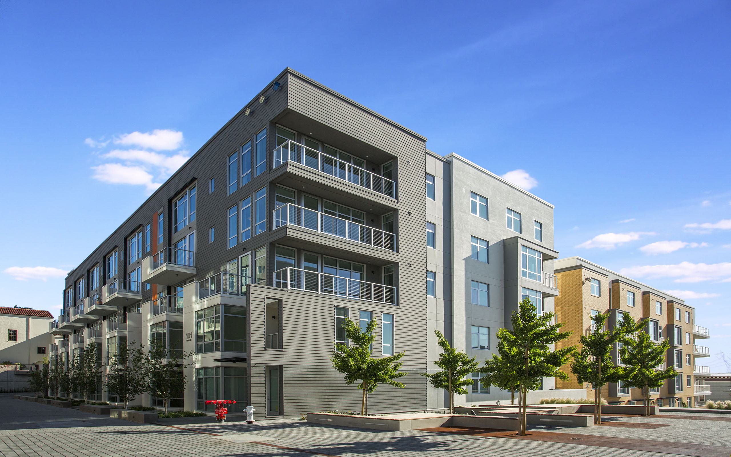 Alchemy contemporary apartment building in Hayes Valley with clean lines, large windows, and private balconies, framed by sleek siding and light neutral tones. Young trees line the paved courtyard below, giving the space a modern, walkable neighborhood feel.