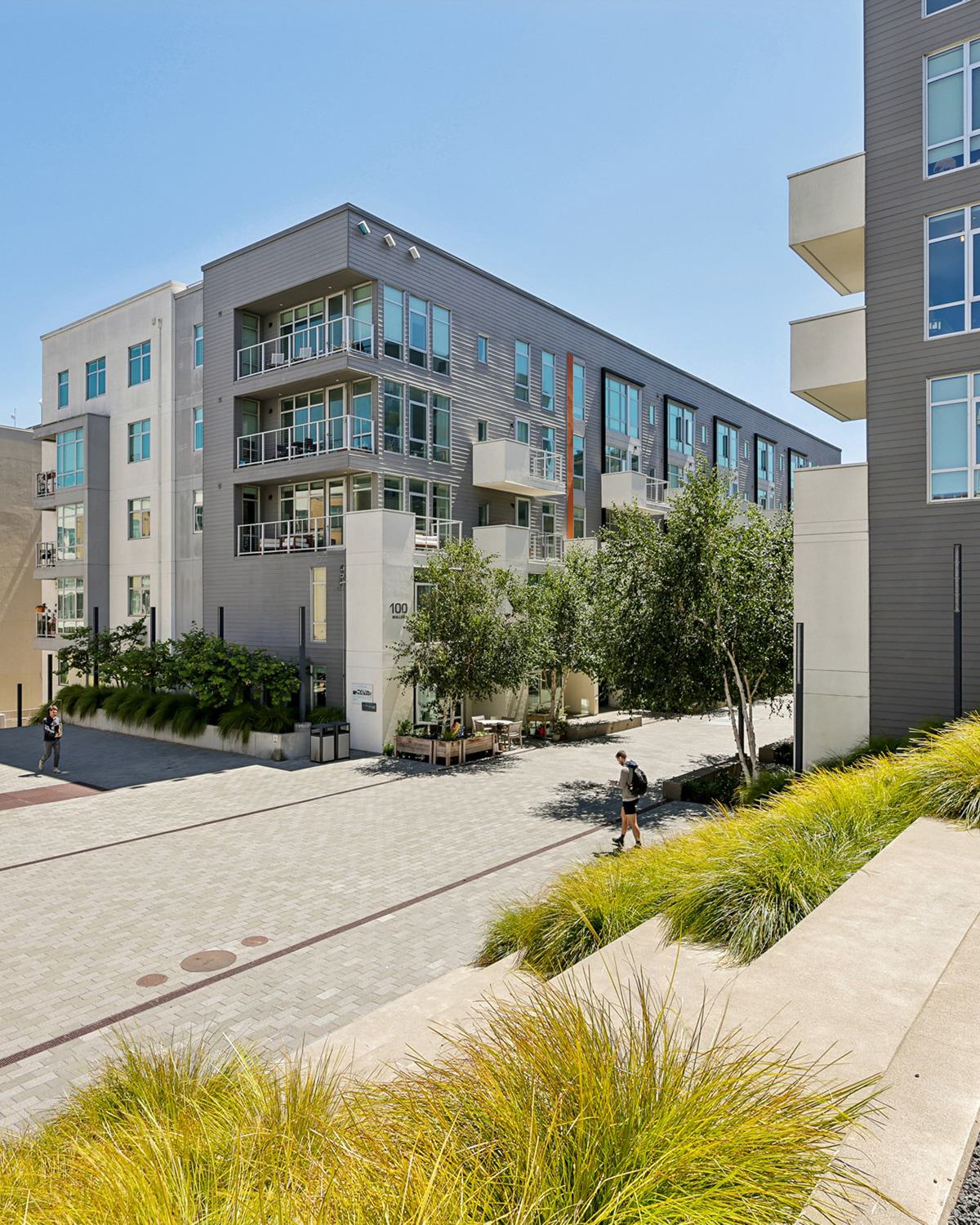 Alchemy modern apartment building with large windows and balconies surround a tree-lined pedestrian courtyard with landscaped planters, steps, and people walking in the sun.