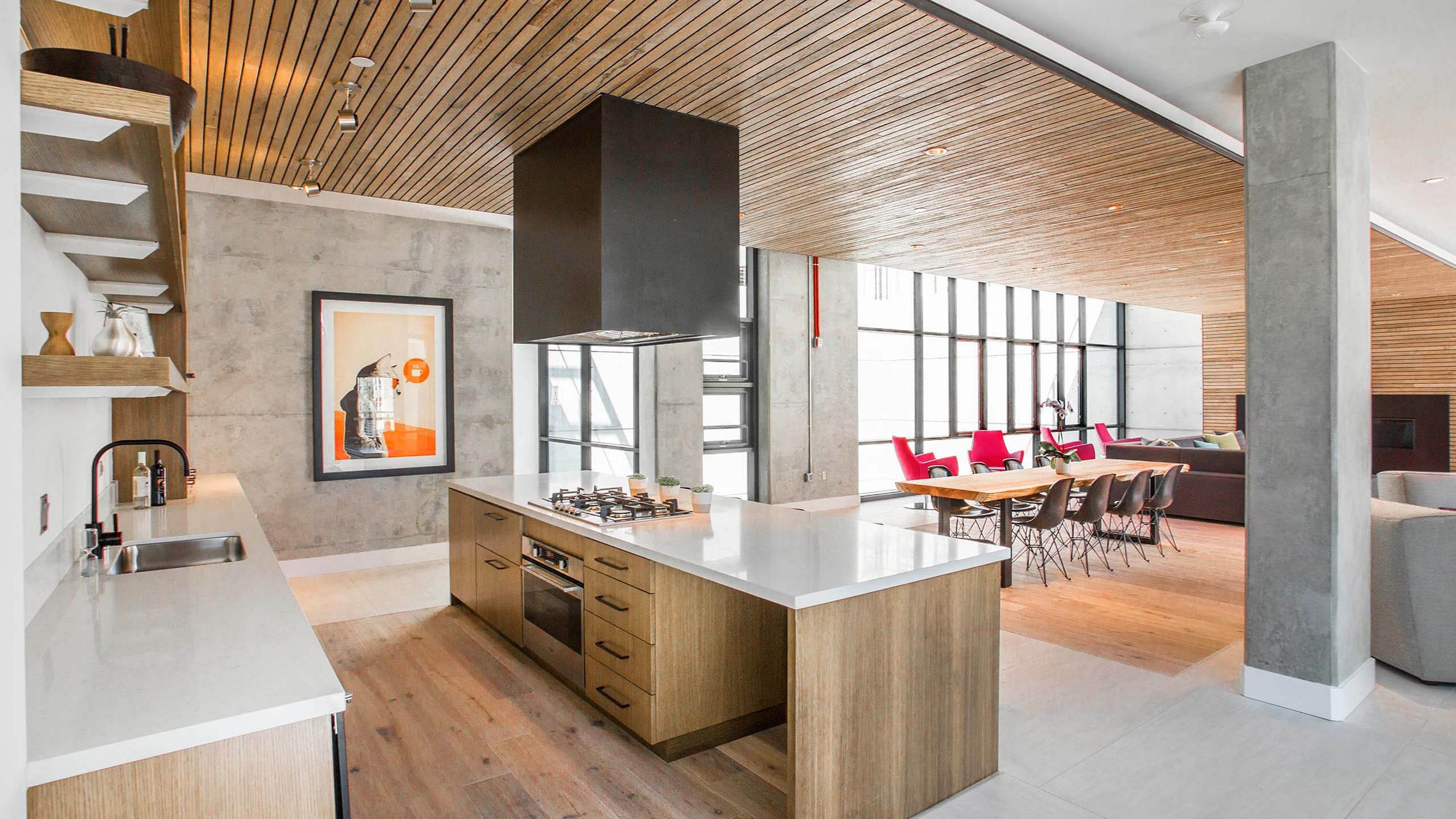 Bright, modern community kitchen and lounge area with sleek finishes, wood accents, a large island with cooktop, communal dining table, and cozy seating under slatted wood ceilings.