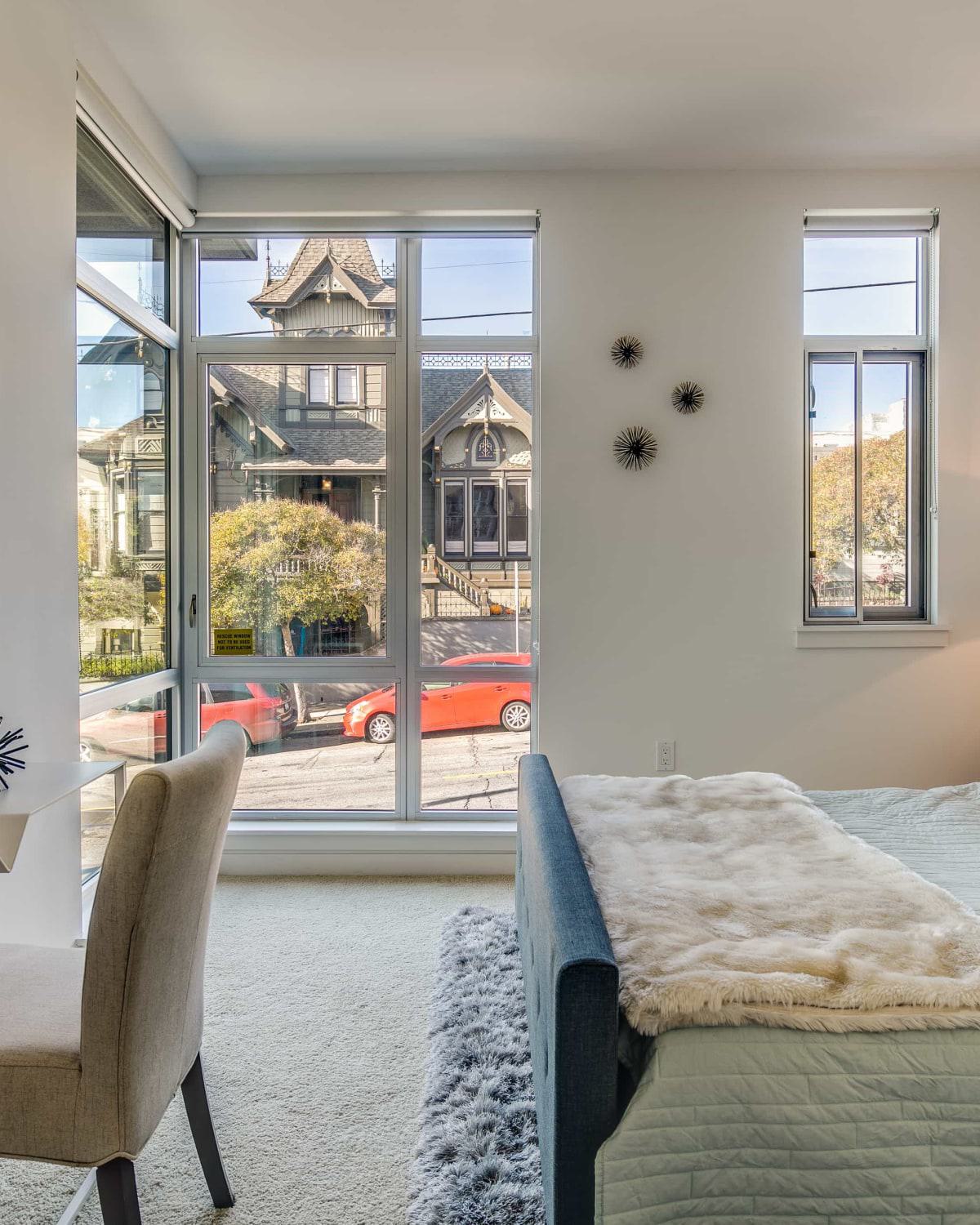 Bright, modern bedroom with large windows overlooking a historic Victorian home, featuring a cozy bed, desk, contemporary decor, and natural light streaming through a floor-to-ceiling window.