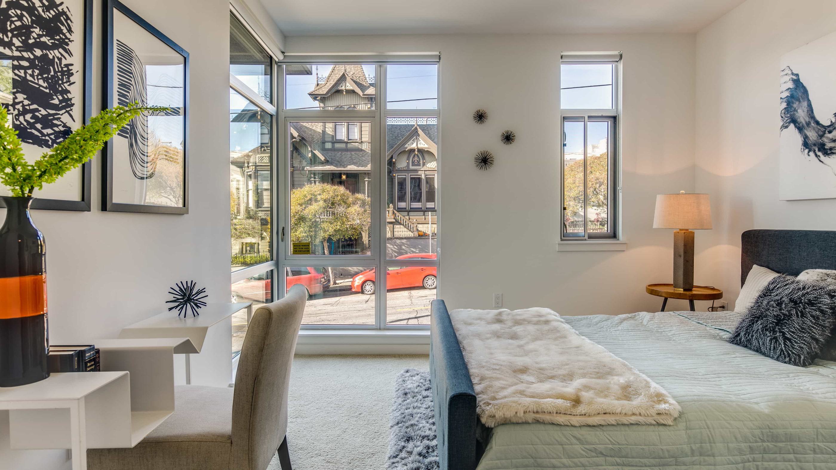 Bright, modern bedroom with large windows overlooking a historic Victorian home, featuring a cozy bed, desk, contemporary decor, and natural light streaming through a floor-to-ceiling window.