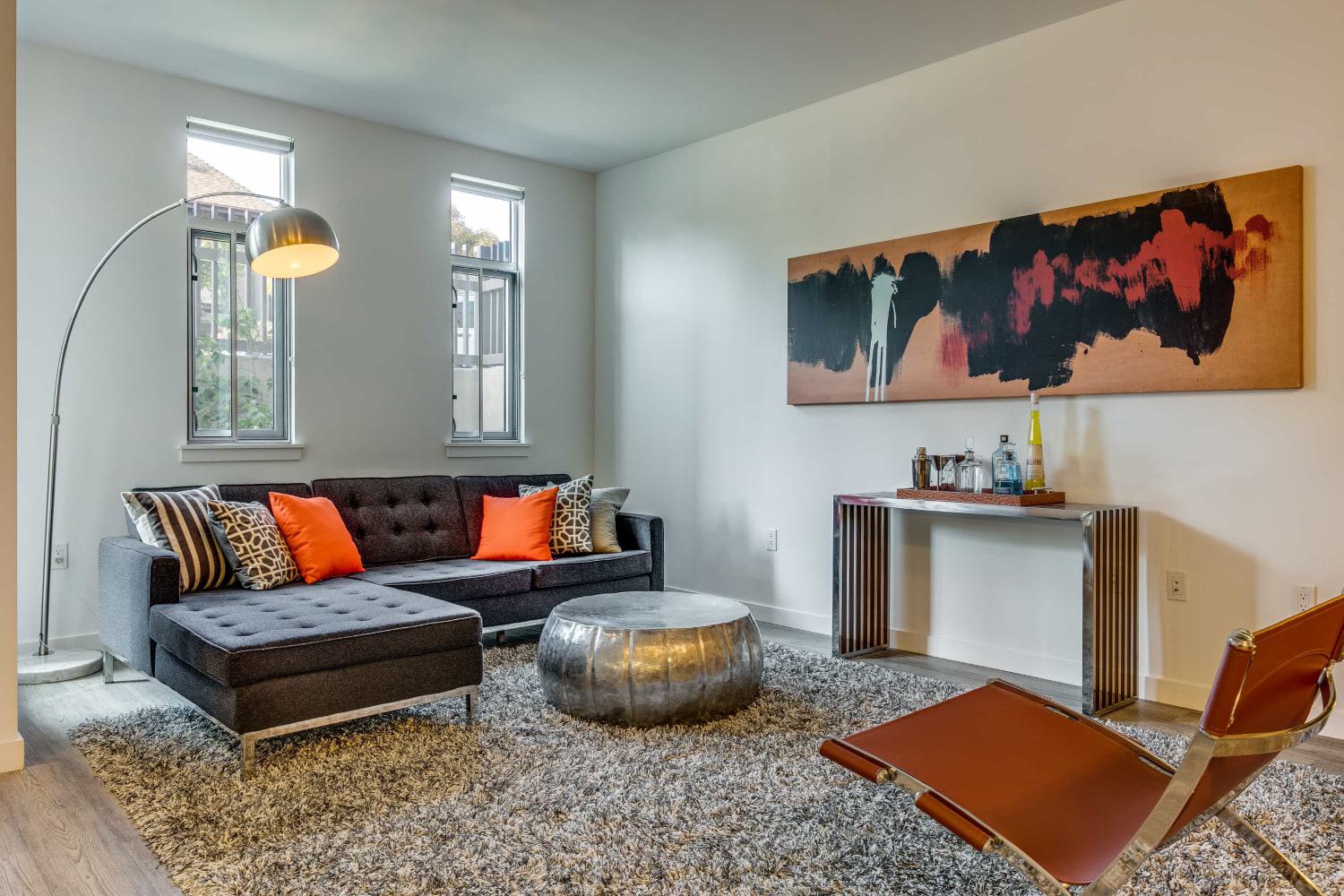 Modern living room with a dark sectional sofa, orange accent pillows, a silver coffee table, abstract wall art, and floor lamp, set on a textured rug beneath tall windows.