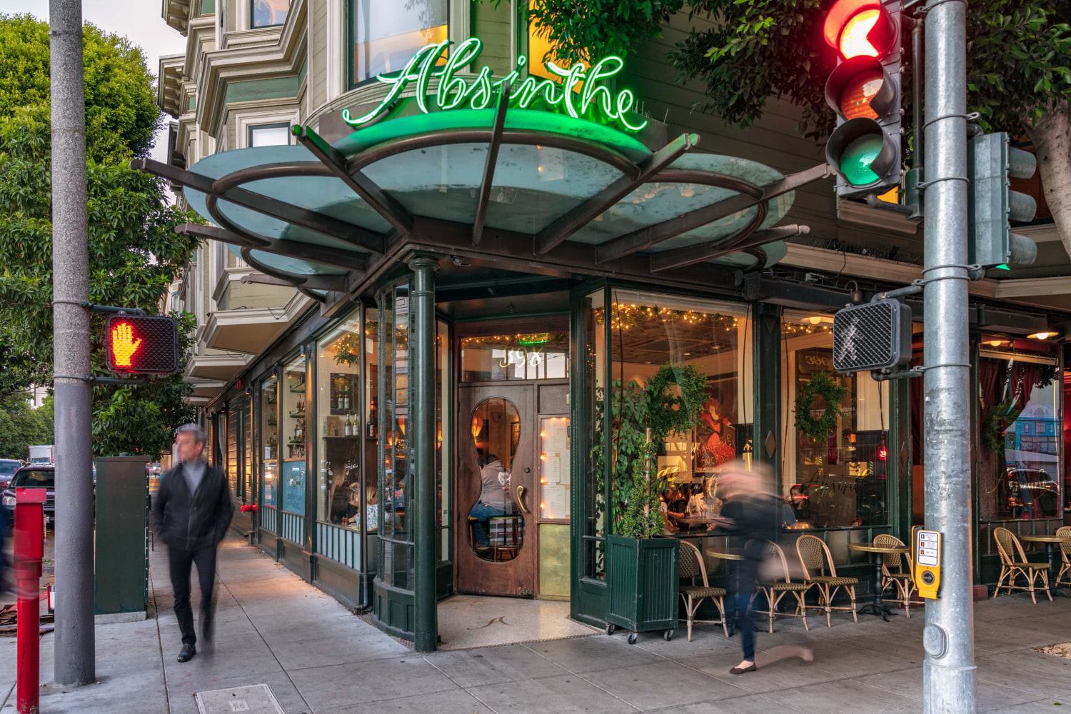 Street view of Absinthe Brasserie & Bar in Hayes Valley, featuring a glowing green neon sign, glass-paneled exterior, and a cozy, warmly lit interior visible through large windows.