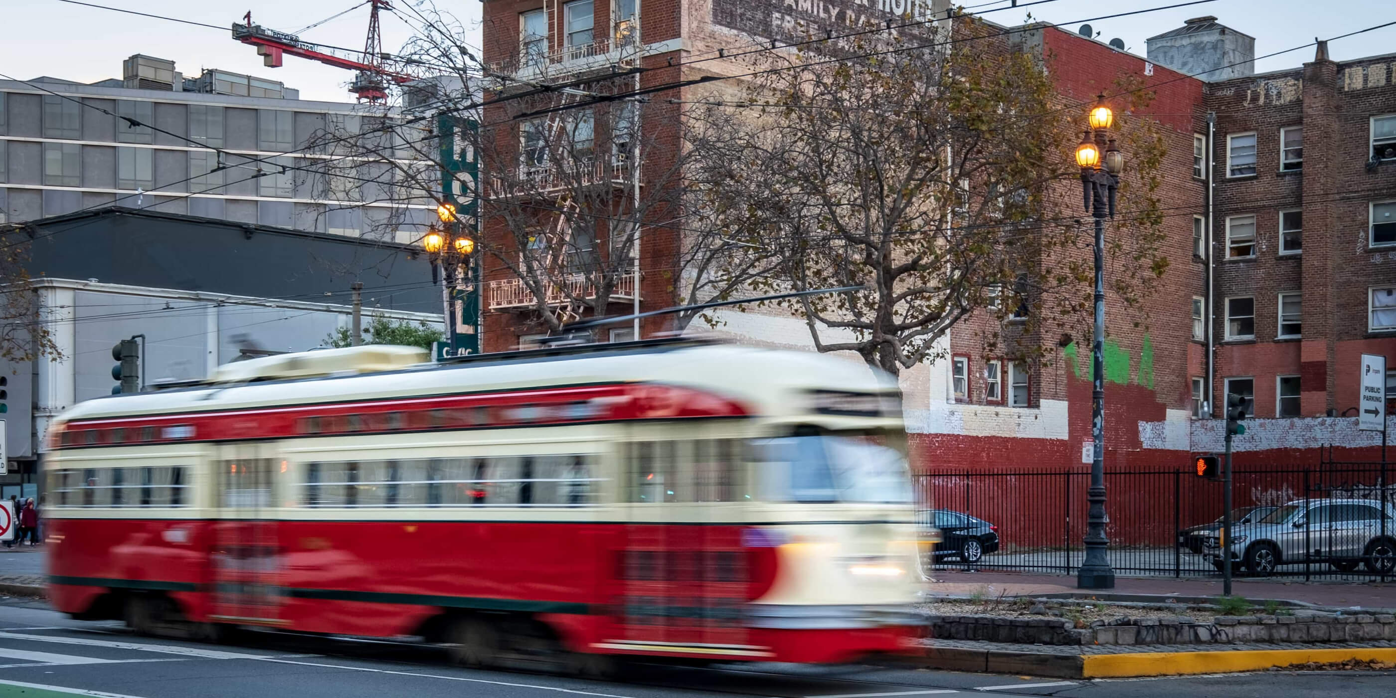 A vintage red and cream PCC streetcar from San Francisco’s F Market & Wharves line moves through Hayes Valley. The scene features brick apartment buildings with fire escapes, a faded wall advertisement, bare winter trees, and classic streetlamps. Overhead electric wires and a construction crane in the background hint at the neighborhood’s historic character and ongoing development.