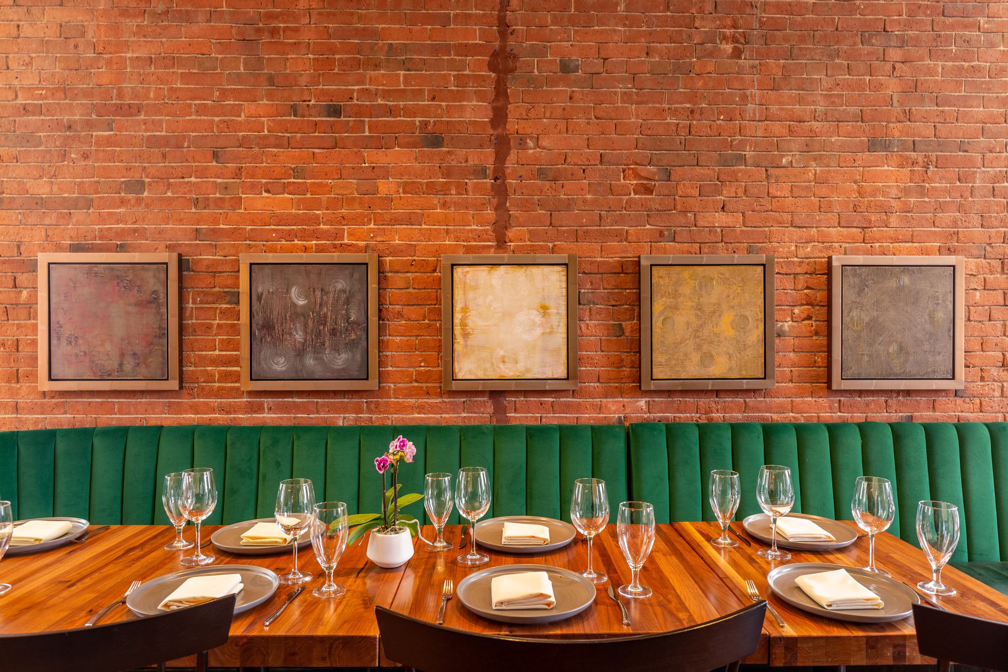 CLINK. restaurant interior with green velvet seating, exposed brick wall, abstract artwork, and a neatly set wooden dining table with glassware and napkins.