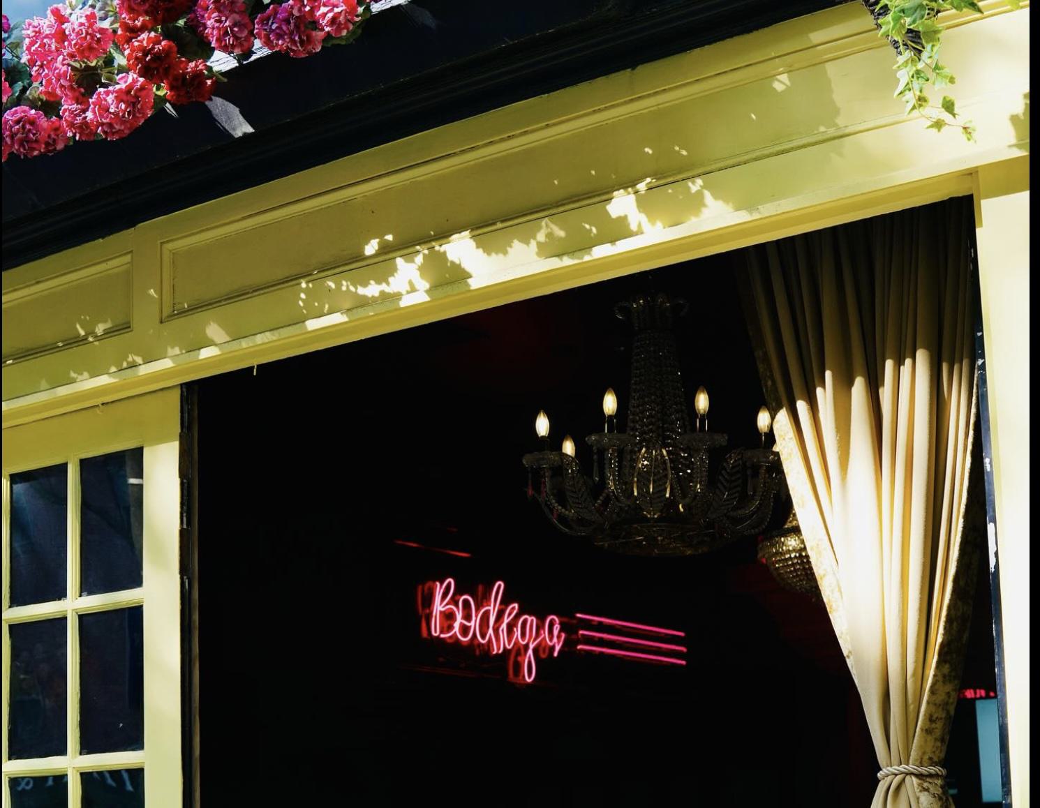 Bodega Canal restaurant entrance with yellow trim, pink flowers, velvet curtains, a crystal chandelier, and a neon “Bodega” sign glowing inside.
