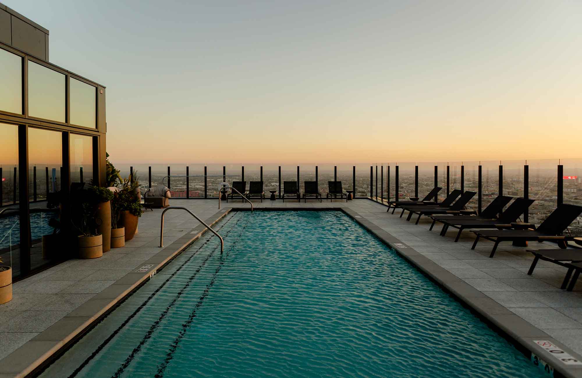 Rooftop pool at Alloy Apartments with city views at sunset, surrounded by lounge chairs and glass safety railing overlooking the Los Angeles skyline.