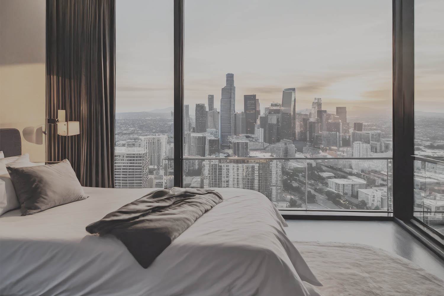 Penthouse bedroom at Alloy featuring a plush bed, dramatic floor-to-ceiling windows, and an expansive balcony with unobstructed views of the downtown Los Angeles skyline at sunset.