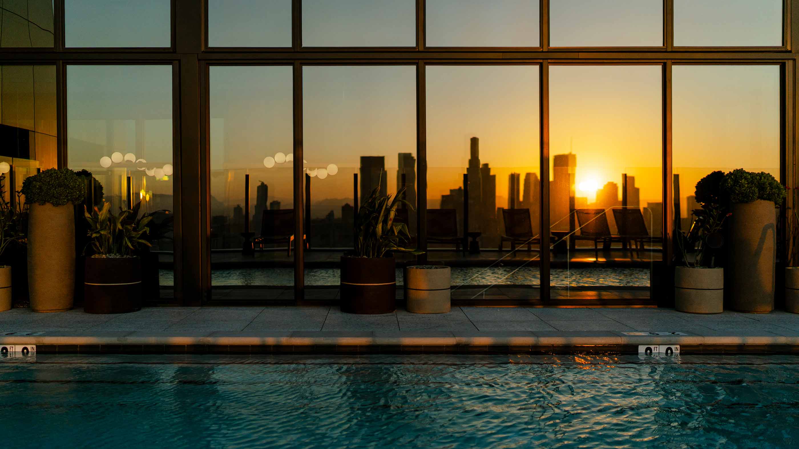 Rooftop pool at Alloy Apartments reflecting the downtown Los Angeles skyline and sunset through large glass windows, with potted plants and lounge chairs visible.