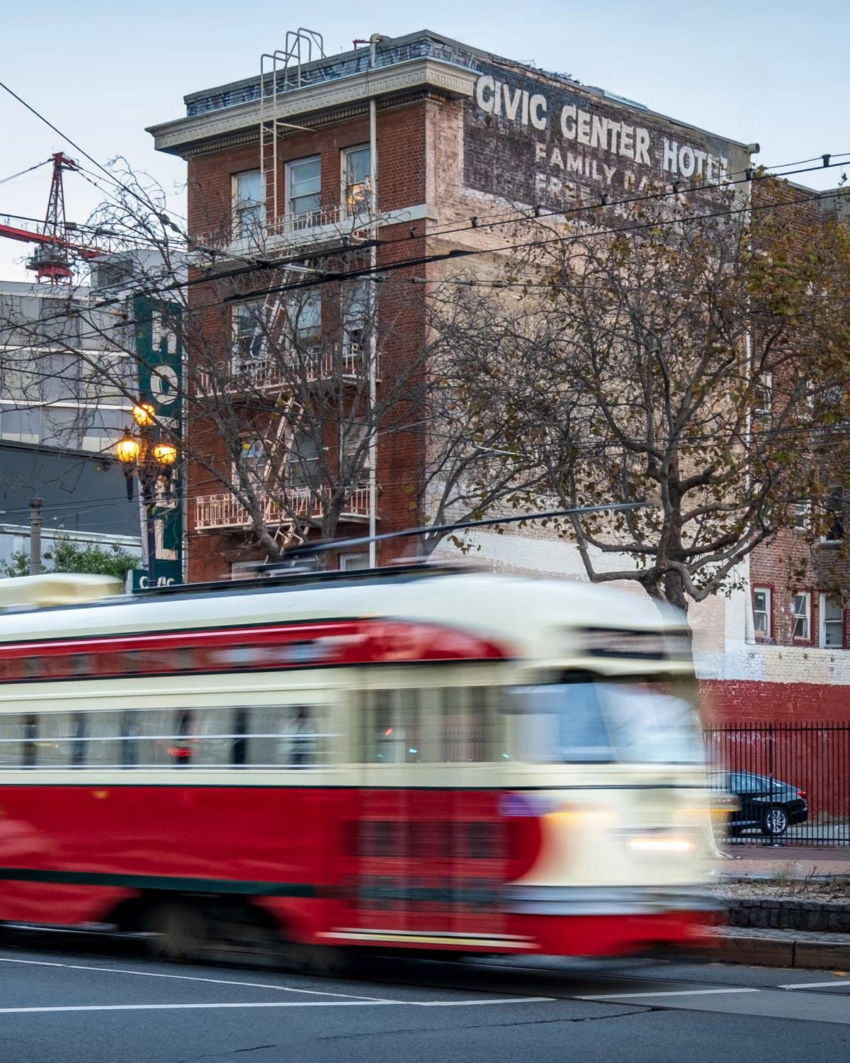 Red and cream Muni streetcar passing a historic brick building with a faded “Civic Center Hotel” sign in San Francisco’, framed by overhead trolley wires and leafless street trees at dusk.
