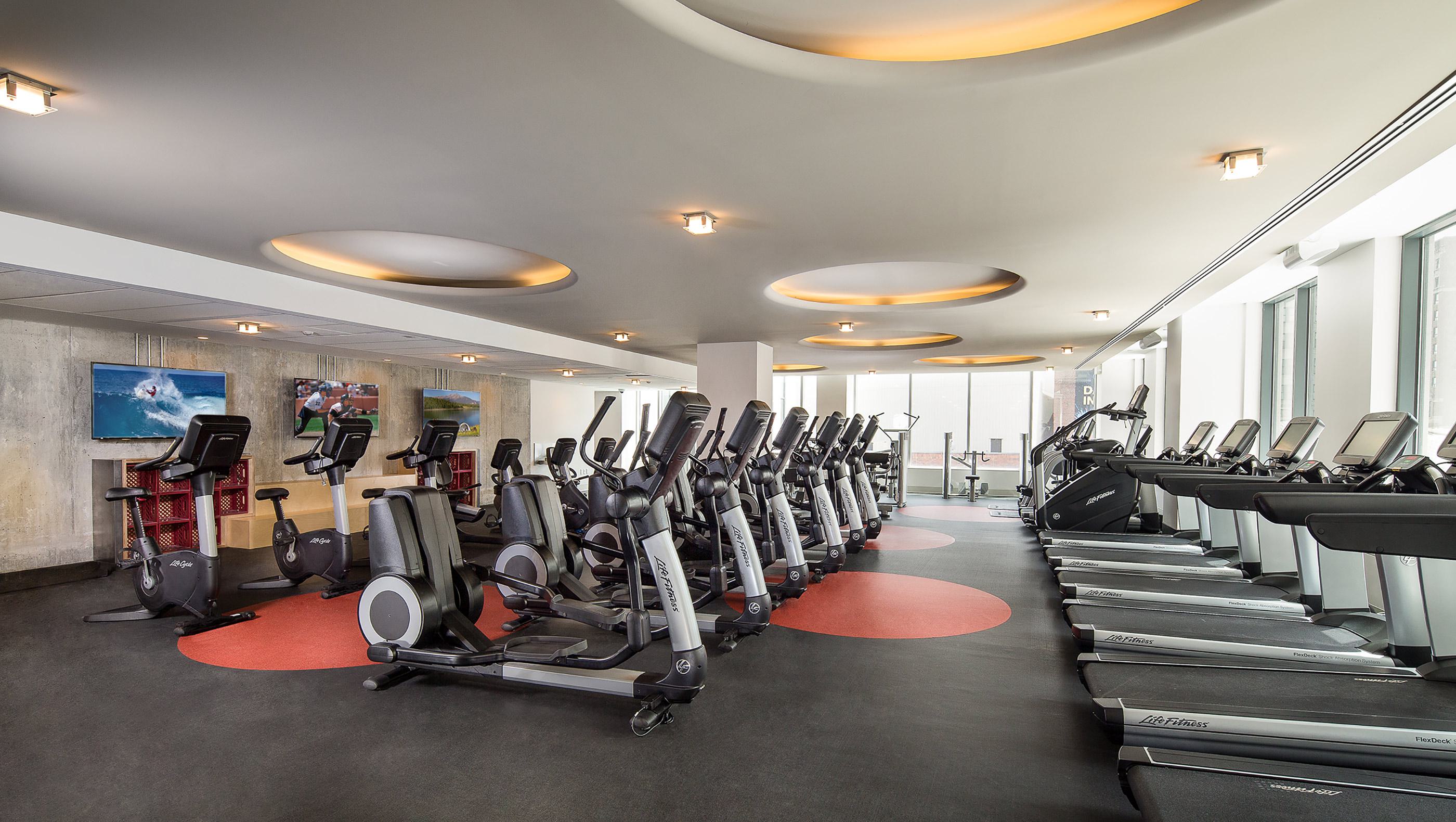 Modern fitness center with rows of Life Fitness treadmills, elliptical machines, and stationary bikes, positioned under circular recessed lighting. Floor-to-ceiling windows line one side of the room, and multiple flat-screen TVs display sports and scenic imagery along a concrete accent wall.