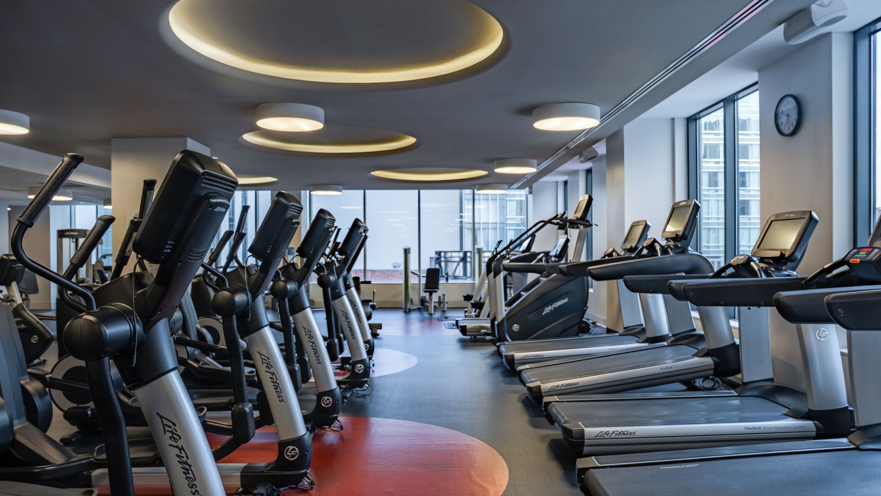 Fitness center with rows of Life Fitness treadmills, elliptical machines, and stationary bikes, positioned under circular recessed lighting. Floor-to-ceiling windows line one side of the room, and multiple flat-screen TVs display sports and scenic imagery along a concrete accent wall.
