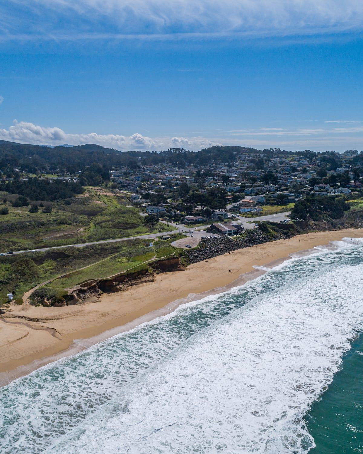 Aerial view of the California coastline near Burlingame, showing sandy beach, ocean waves rolling onto shore, grassy bluffs, and a nearby coastal town under a bright blue sky.
