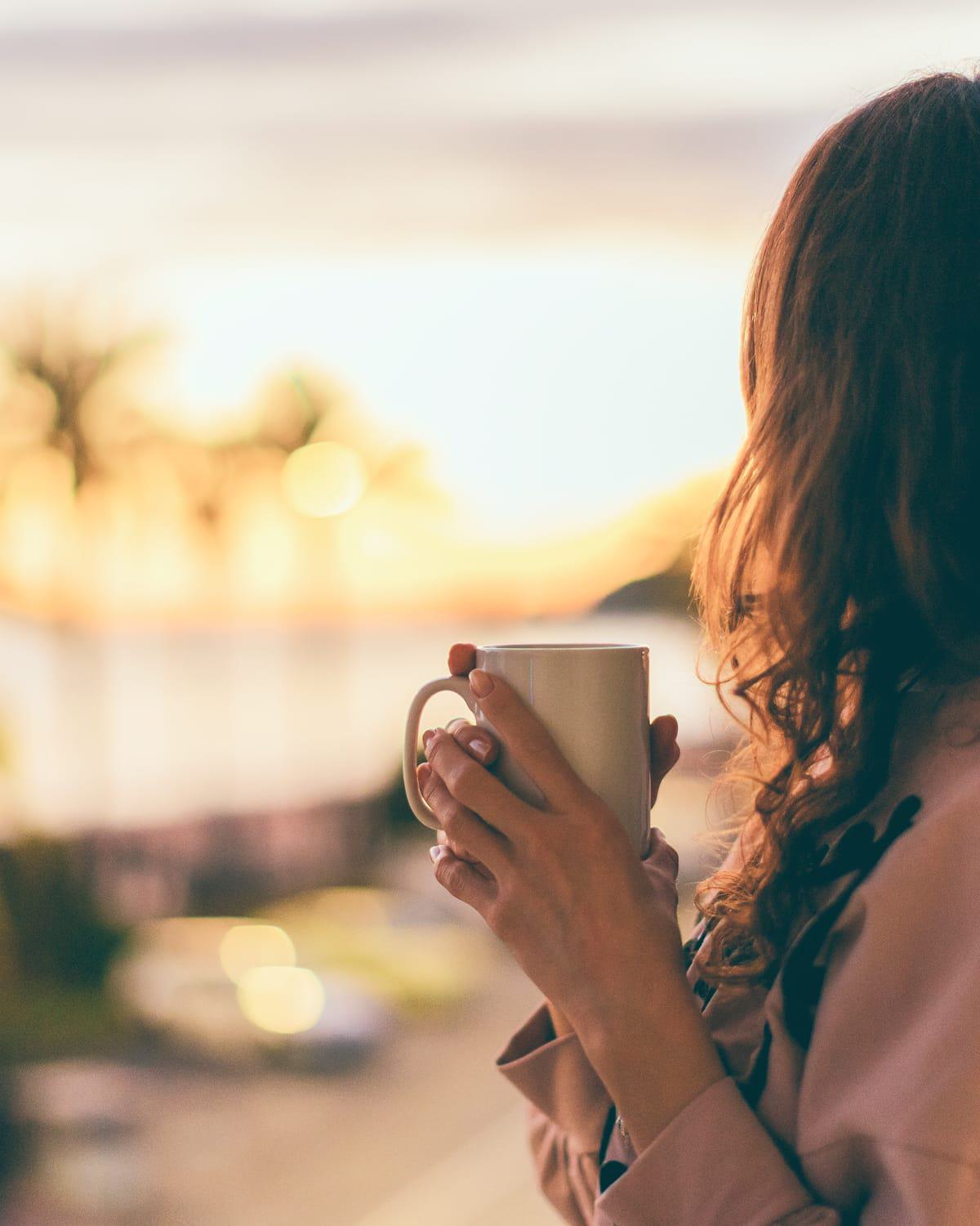 A woman with long, wavy hair stands on a balcony at sunset, holding a mug with both hands. The warm, golden light softens the scene as palm trees and blurred city lights create a tranquil, dreamy backdrop.