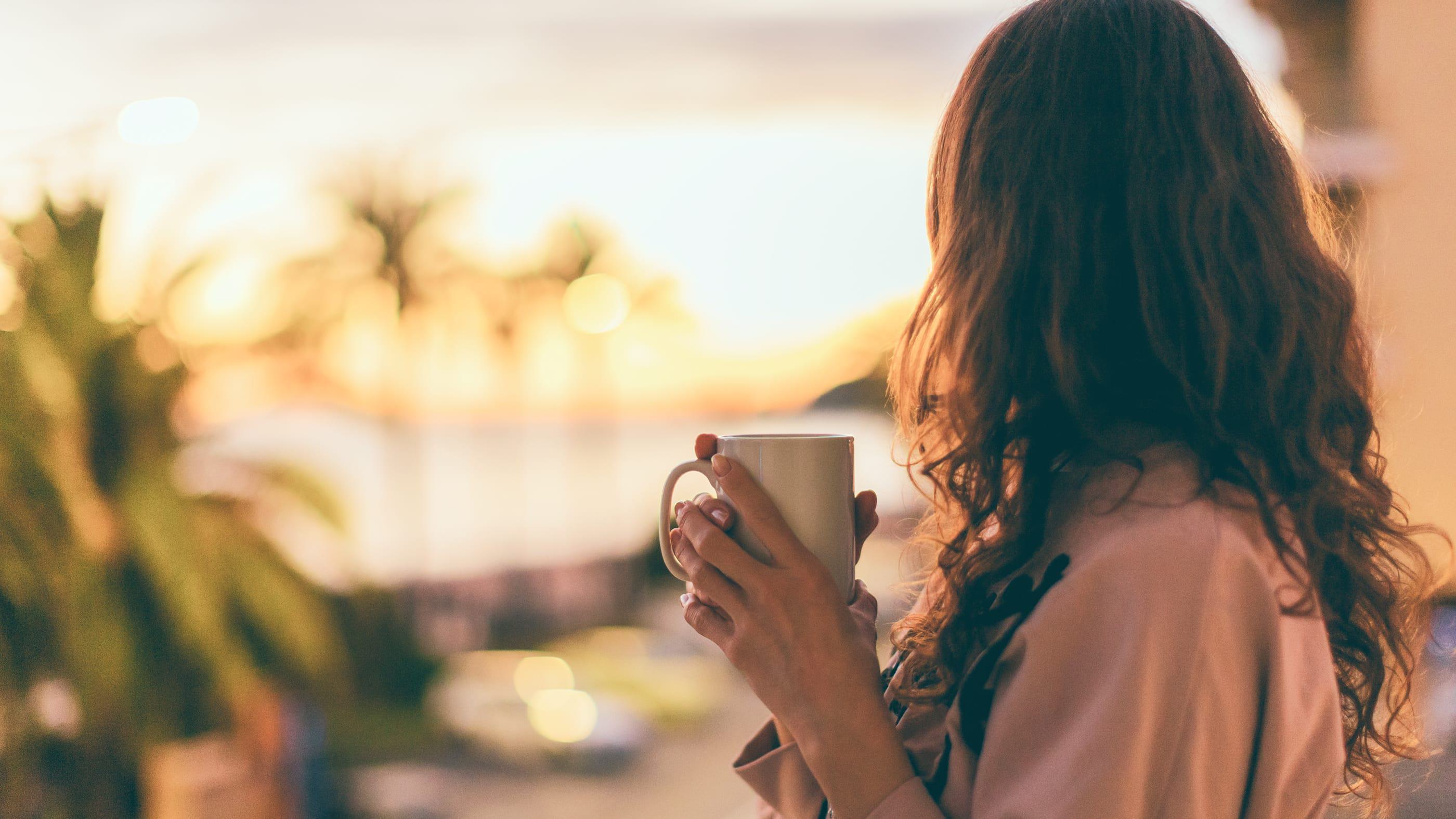 A woman with long, wavy hair stands on a balcony at sunset, holding a mug with both hands. The warm, golden light softens the scene as palm trees and blurred city lights create a tranquil, dreamy backdrop.