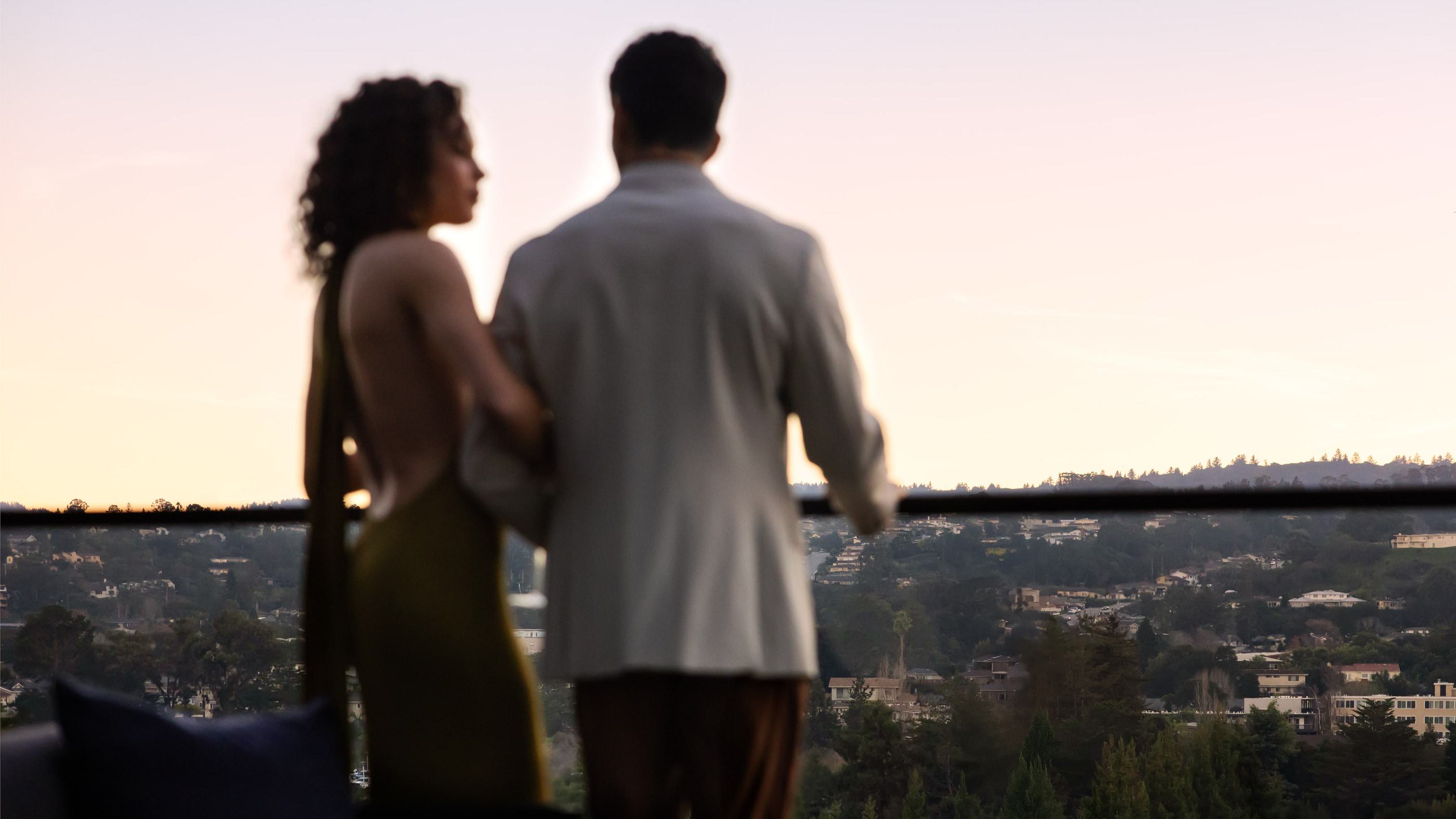 A woman and man stand on a balcony at dusk. The warm, golden light softens the scene as palm trees and and a view of Burlingame create a tranquil, dreamy backdrop.