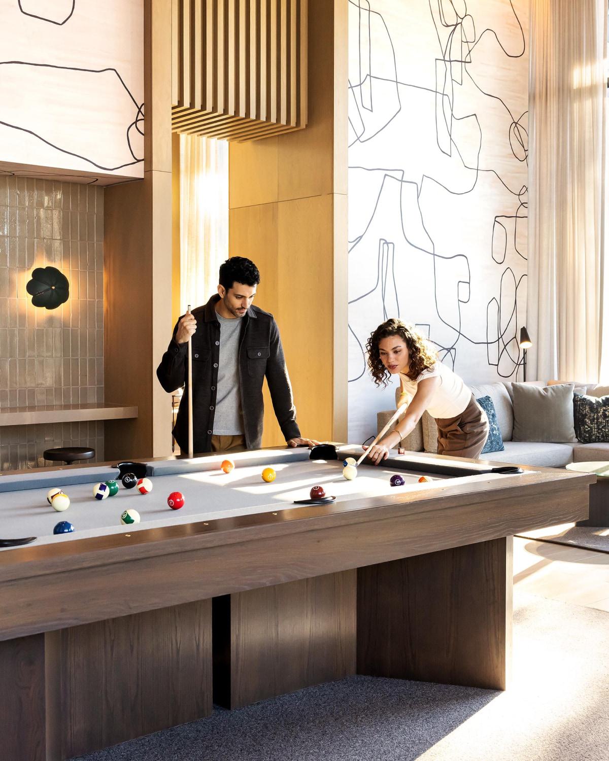 A warm, inviting lounge space with plush seating, a pool table, and abstract lights. Tall windows frame lush greenery and evening views of modern buildings, creating an ideal setting for relaxed social gatherings.