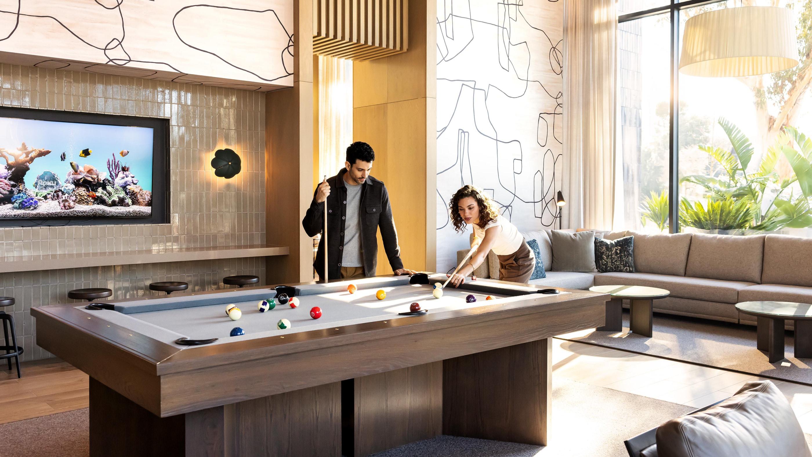 A warm, inviting lounge space with plush seating, a pool table, and abstract lights. Tall windows frame lush greenery and evening views of modern buildings, creating an ideal setting for relaxed social gatherings.