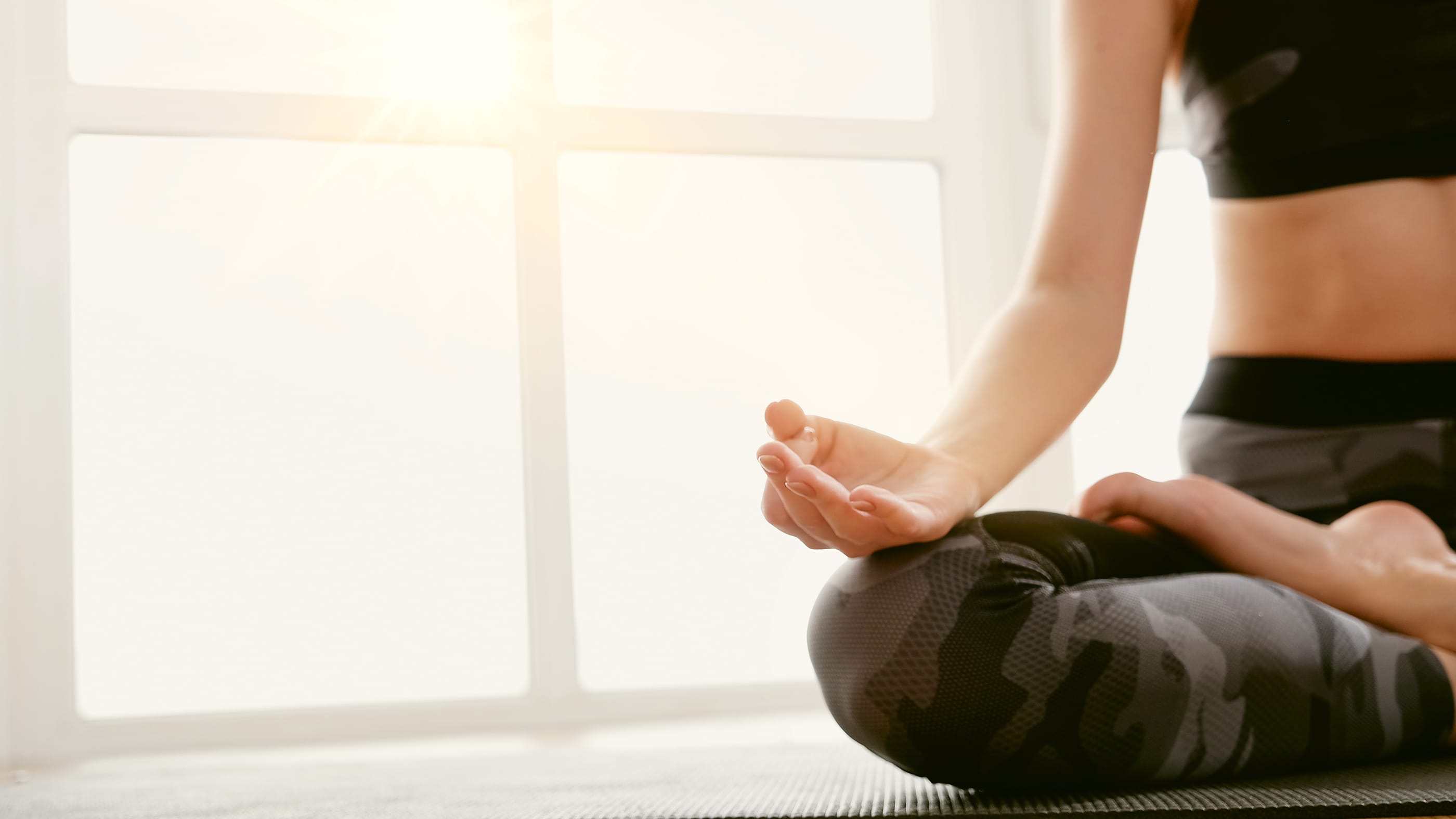Close-up of a person in athletic wear seated in a cross-legged meditation pose on a yoga mat, with morning sunlight streaming through a nearby window. The serene setting highlights calm, focus, and mindful movement.