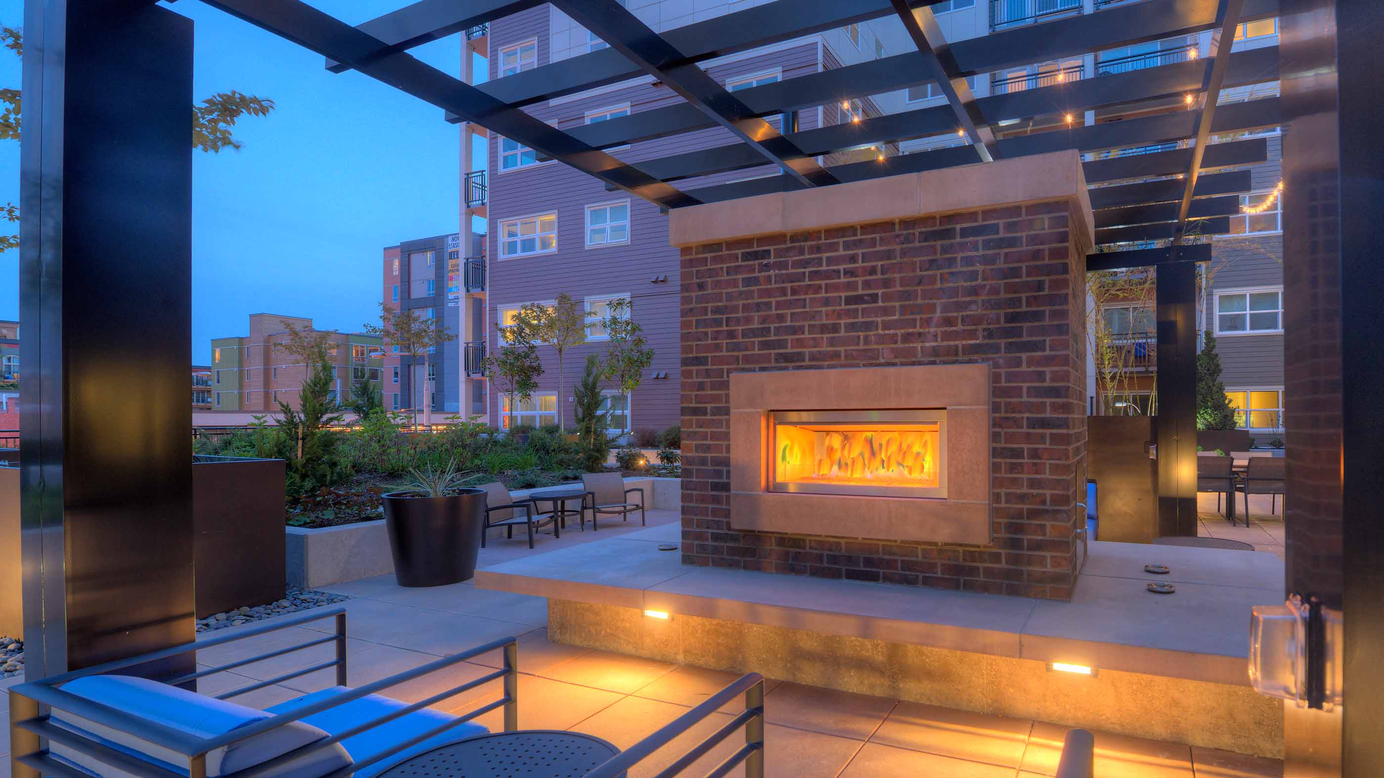 A double-sided brick outdoor fireplace glows warmly under a modern black pergola at dusk, surrounded by seating and courtyard landscaping at Urbana Apartments in Seattle