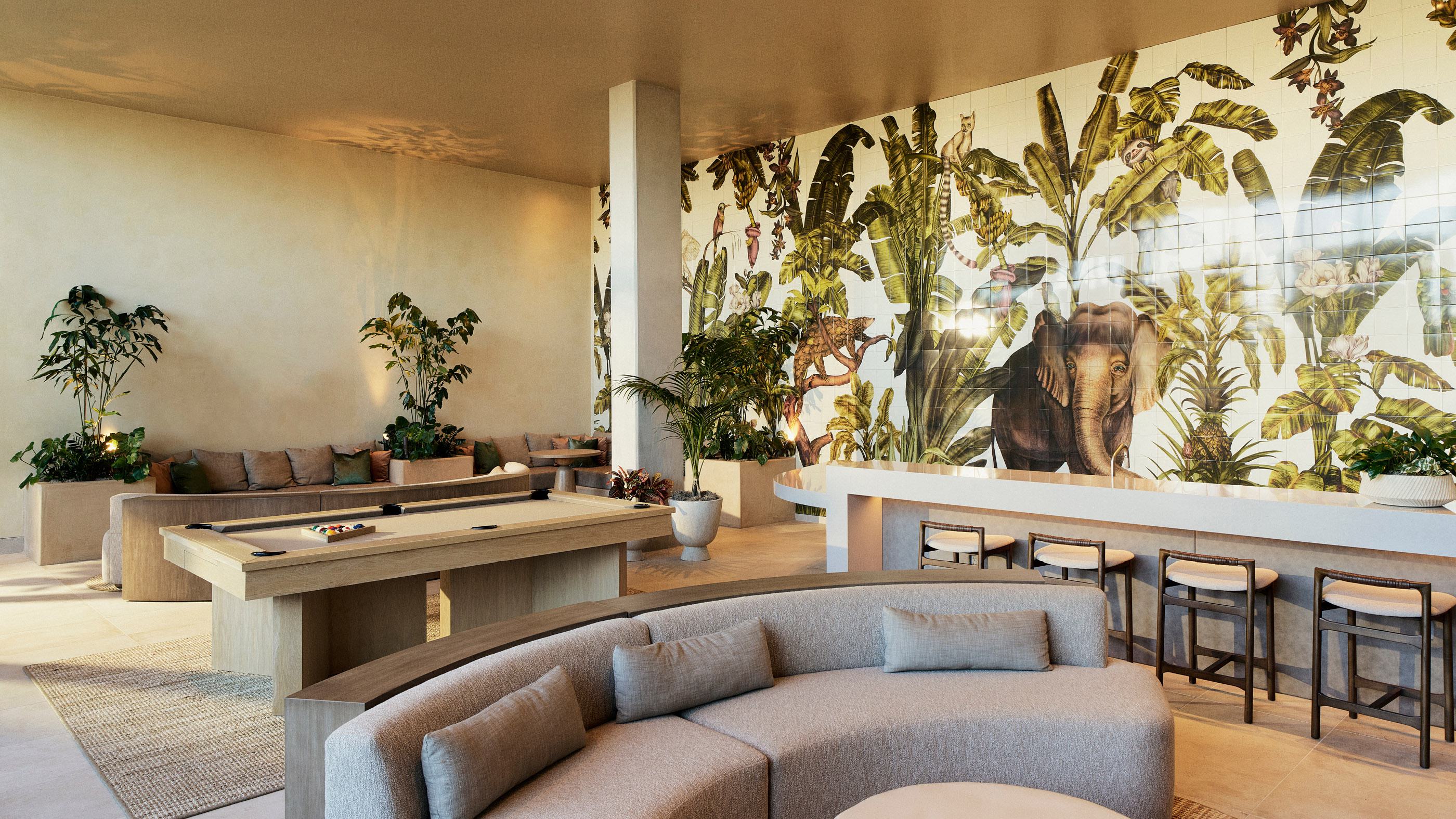 Spacious club lounge featuring a pool table, curved beige sofas, and a large botanical wall mural with tropical plants and an elephant illustration. The room includes a bar area with counter seating, a wall-mounted TV, and warm, natural wood finishes throughout.