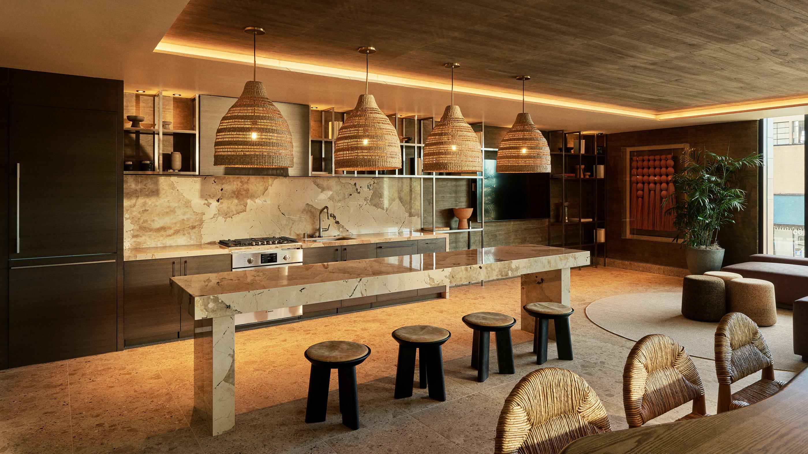 Warm-toned communal dining lounge and kitchen with wood-paneled walls, pendant lighting, long dining tables, open shelving, and a cozy sectional seating area.