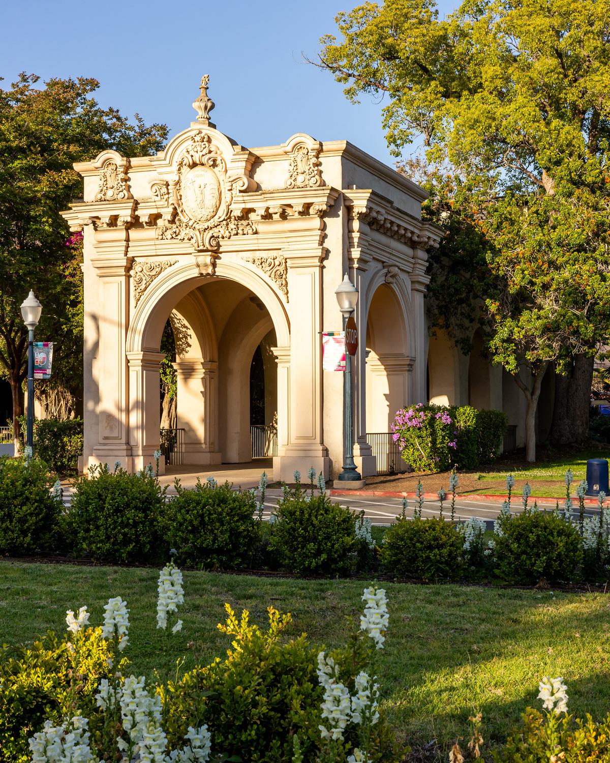 Sunlight hits the ornate Cabrillo Bridge entrance at Balboa Park in San Diego, surrounded by green landscaping and blooming flowers.
